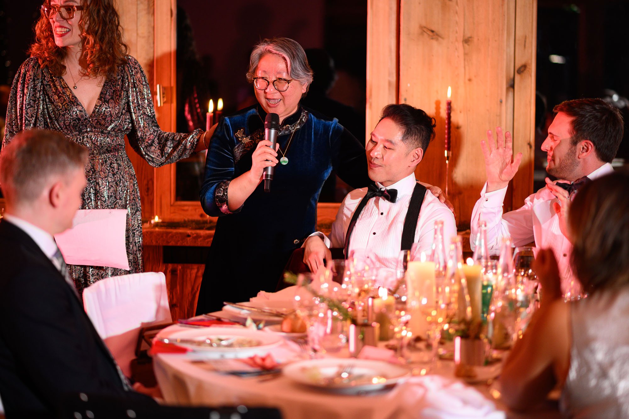 Une femme âgée avec des lunettes parle au micro lors d'un dîner de mariage, entourée de personnes habillées élégamment, avec des décorations de fête et des bougies sur la table.