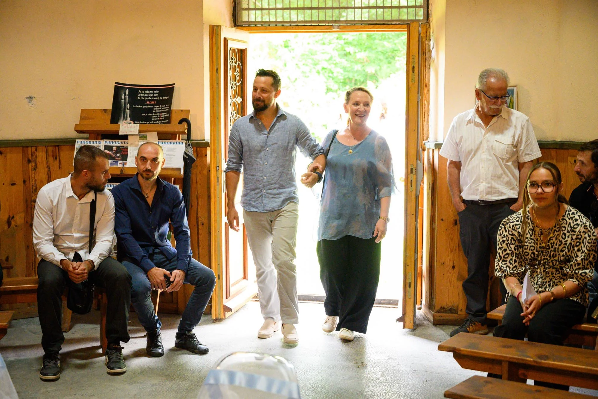 Groupe de personnes entrant dans une pièce lors d'une réunion ou d'un événement social, dans un intérieur en bois avec une porte ouverte laissant passer la lumière naturelle.