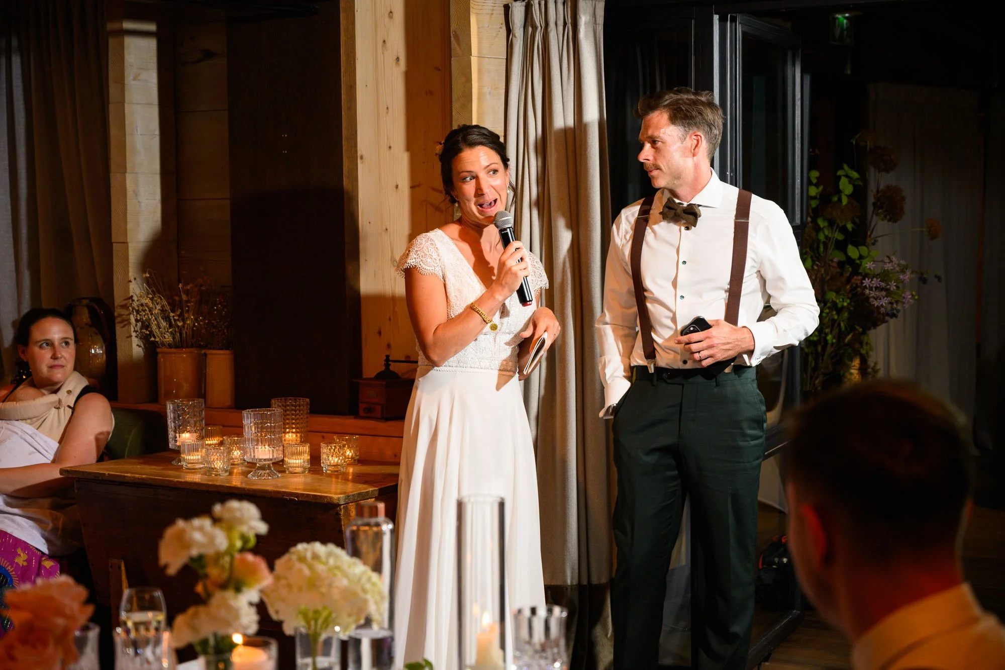 Un mariage avec deux personnes qui parlent lors d'une cérémonie, une femme avec une robe blanche et un homme avec une chemise blanche, cravate noire et bretelles, dans une salle décorée avec des fleurs et des bougies.