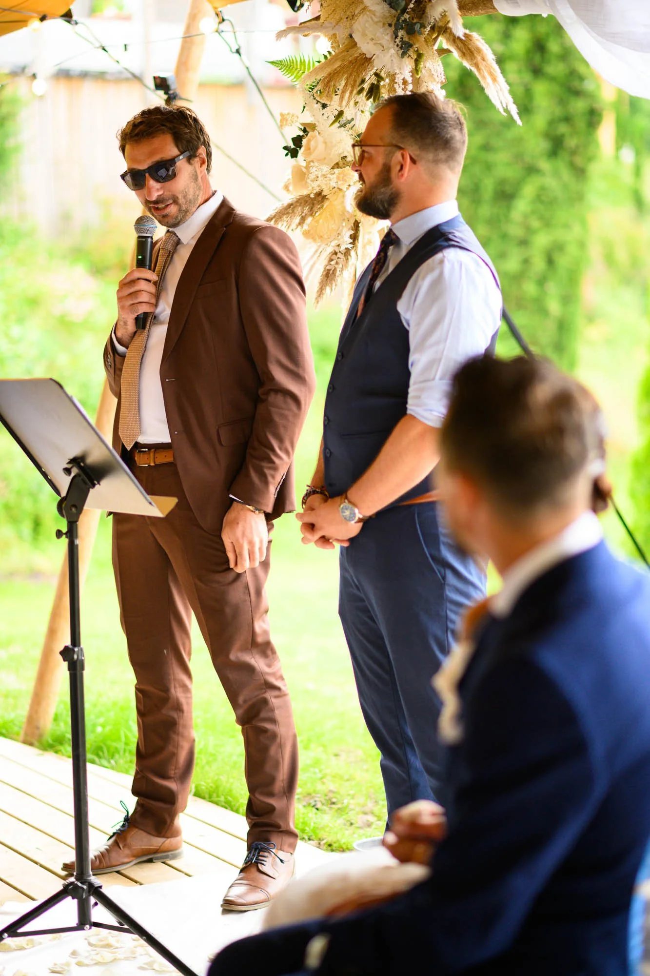 Un homme en costume marron parle dans un micro lors d'une cérémonie en plein air, avec trois autres hommes en costumes, dans un cadre verdoyant avec décoration florale. L'atmosphère semble formelle et festive.