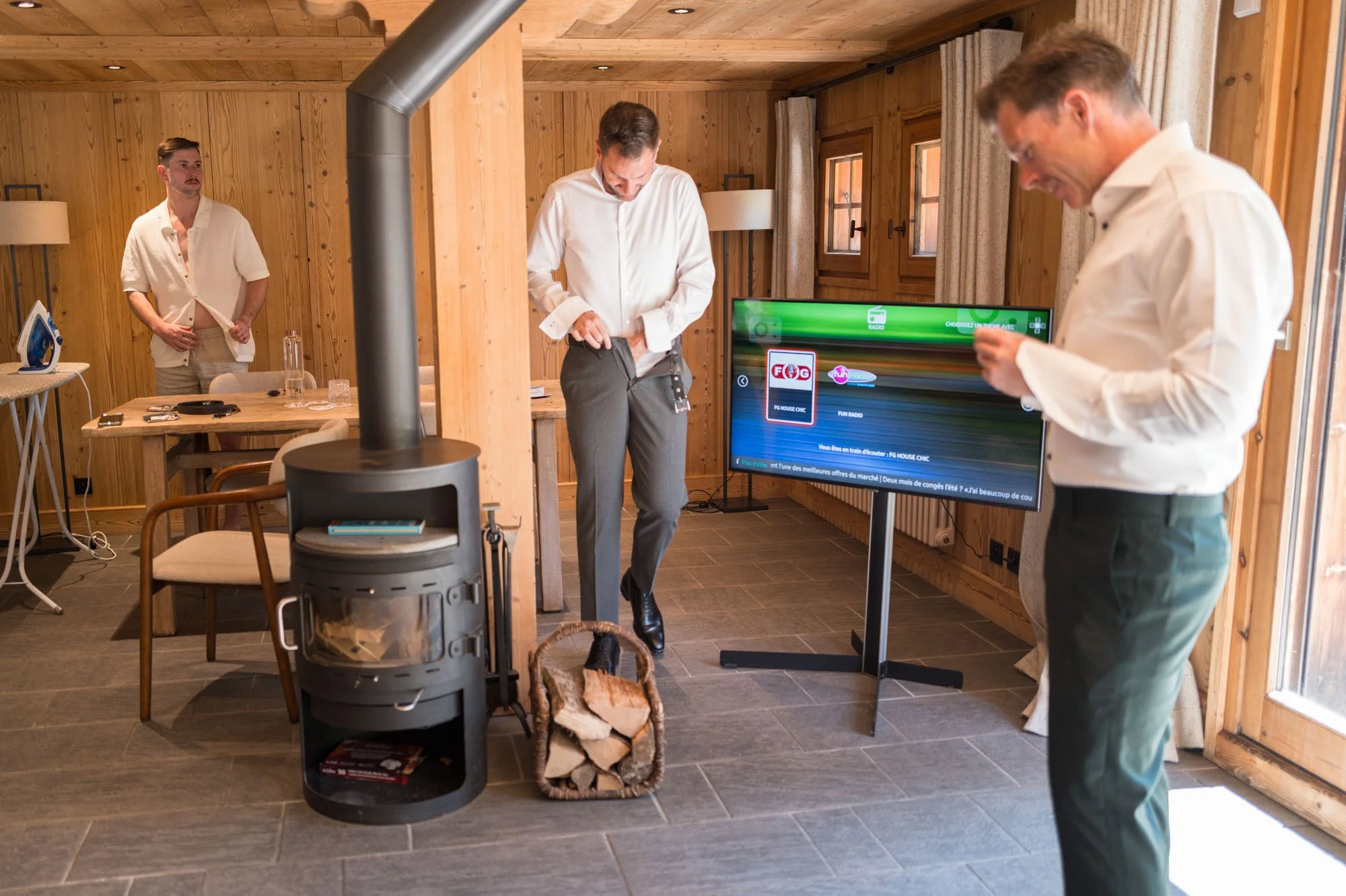 Trois hommes en vêtements décontractés dans une pièce en bois, deux d'entre eux regardent leur téléphone et l'autre ajuste sa poche, avec une cheminée en poêle à bois, un écran TV, et une table avec des objets.