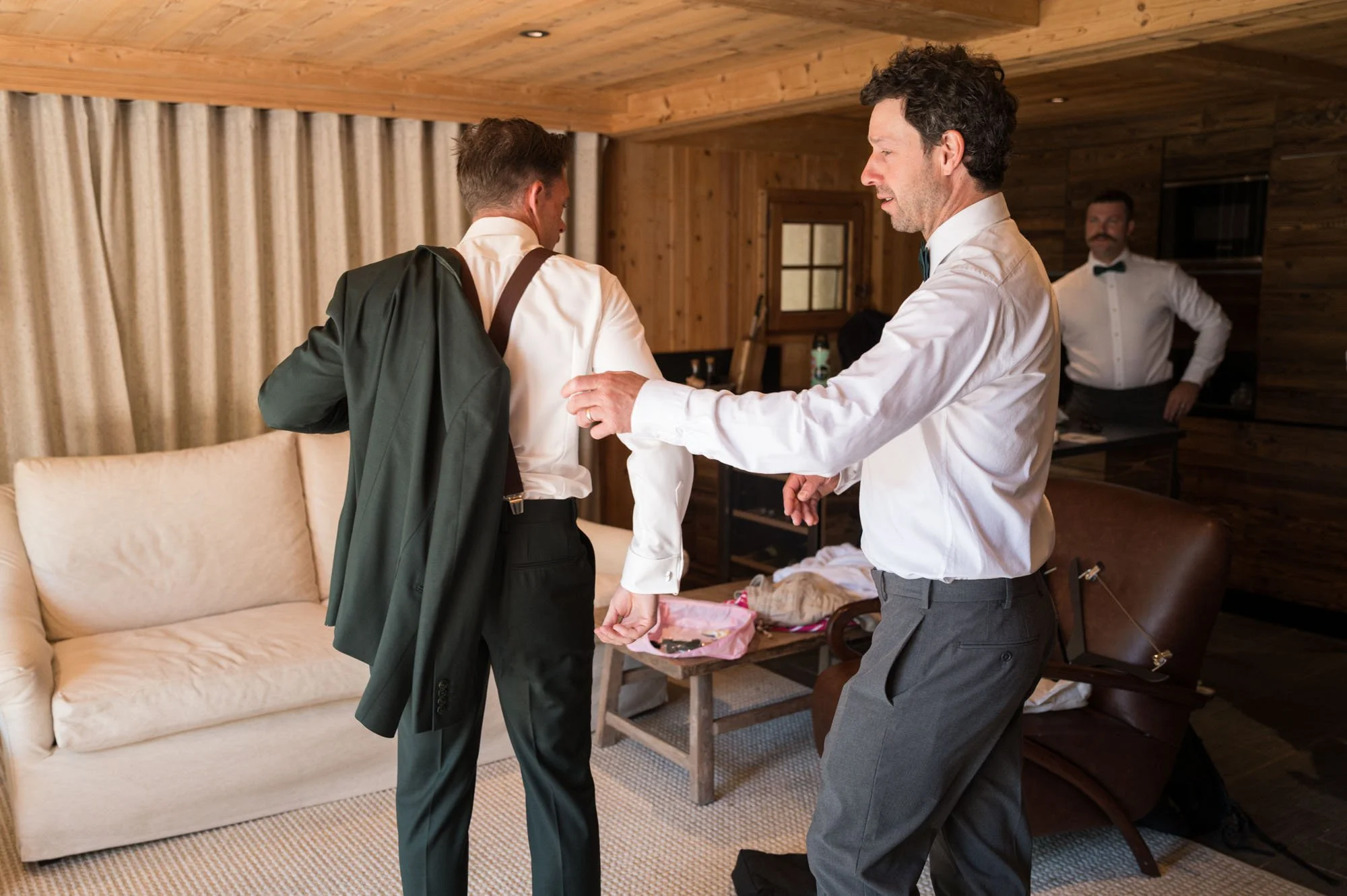 Trois hommes en costumes dans une pièce en bois, l'un aide l'autre à mettre sa veste, un troisième homme en arrière regarde.