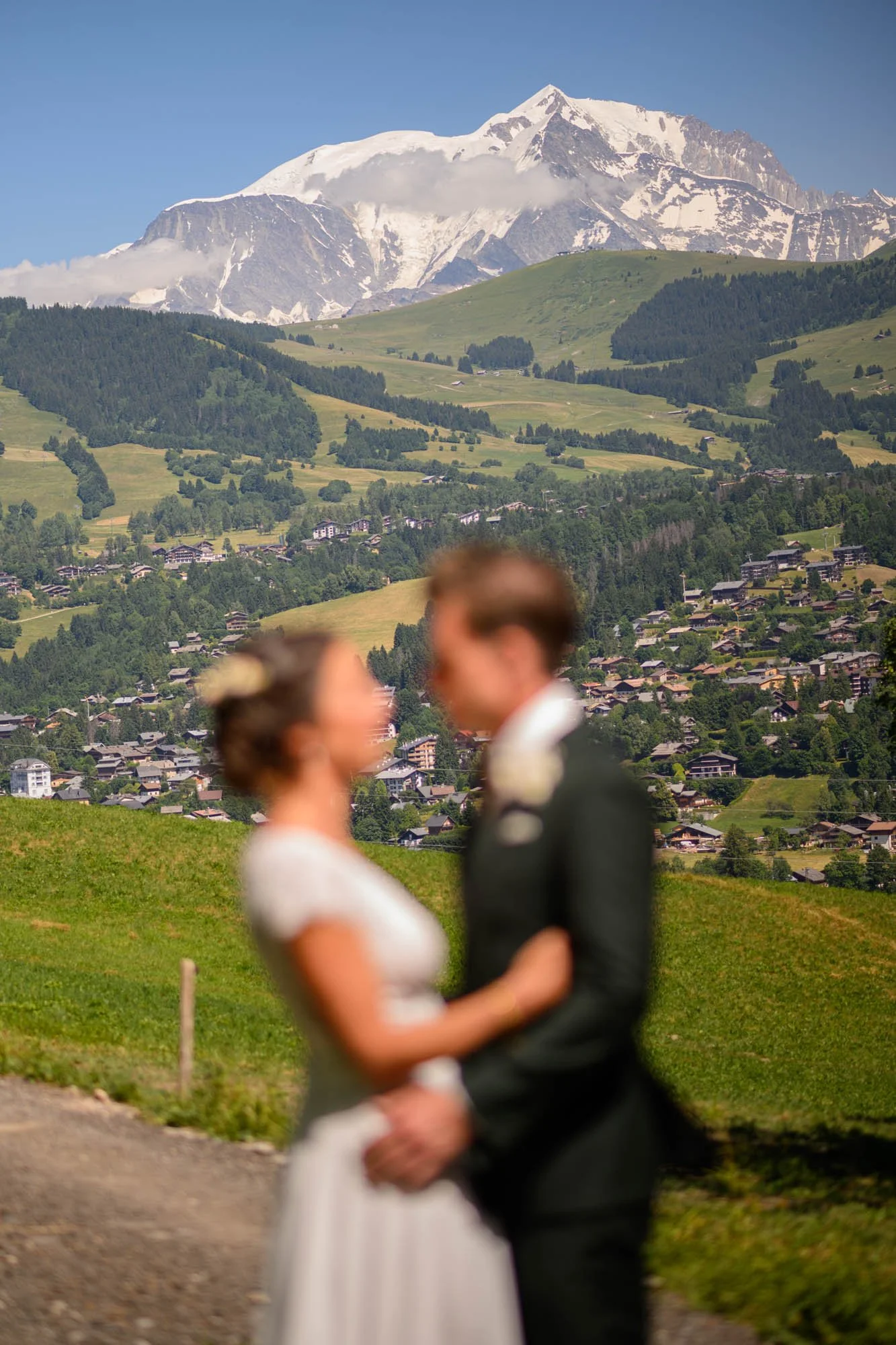 Deux personnes habillées en vêtements de mariage, un homme en costume noir et une femme en robe blanche, se tenant par la taille dans un paysage de montagnes et de prairies vertes avec une ville en contrebas.
