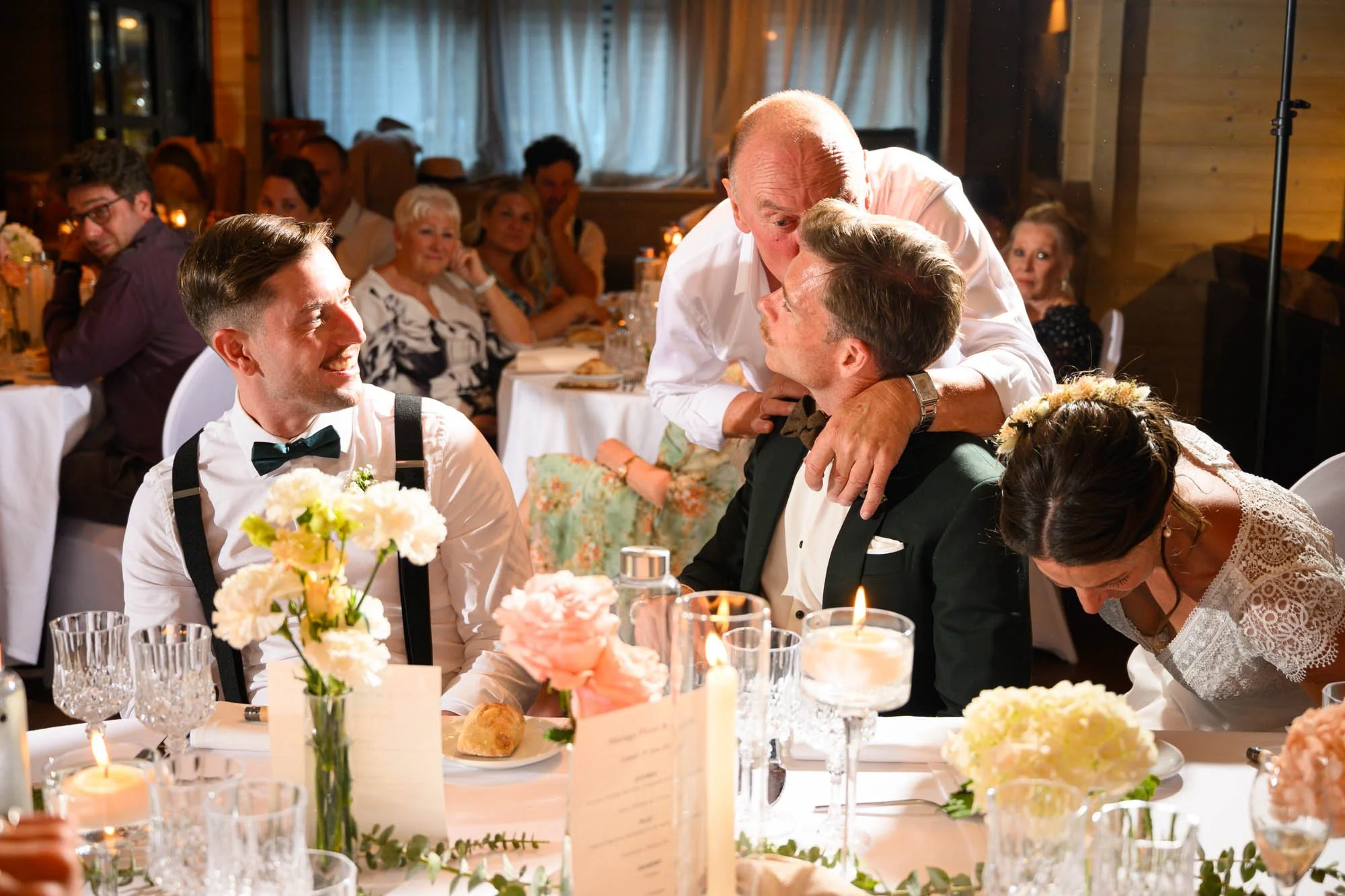 Un groupe de personnes lors d'un mariage, un homme âgé embrasse passionnément un homme en costume, entourés d'invités assis à une table décorée de fleurs et de bougies.