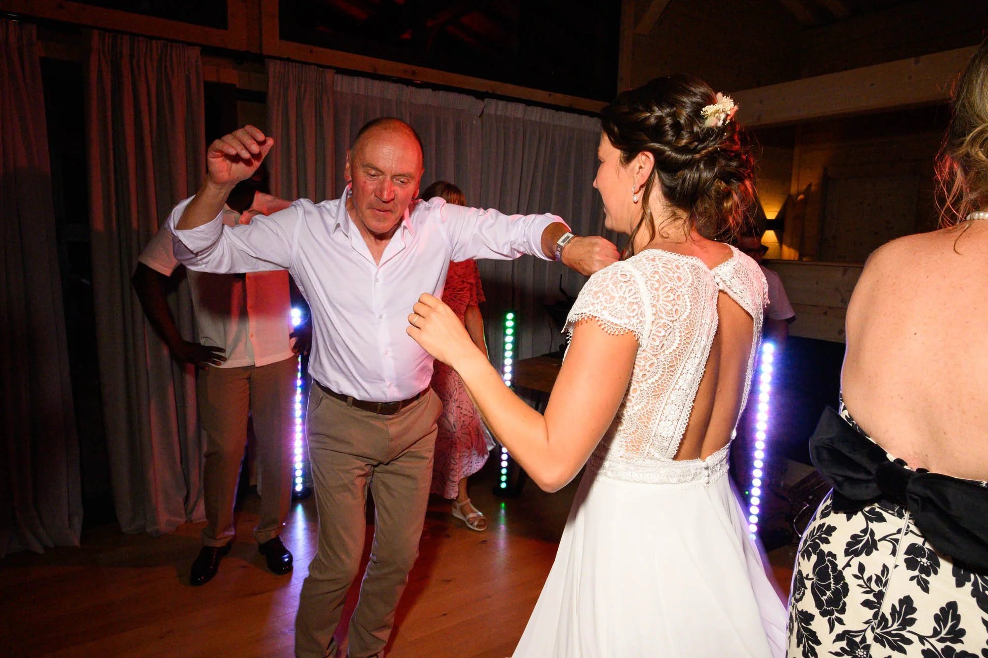 Un homme et une femme dansent lors d'une célébration, probablement un mariage, sous un éclairage coloré avec d'autres personnes en arrière-plan.