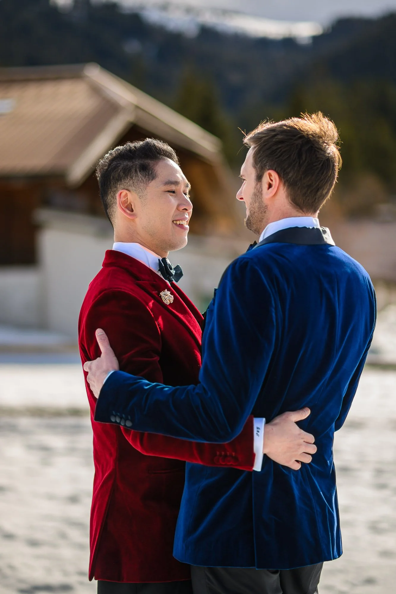 Deux hommes en costumes élégants, se regardant et se tenant dans une embrassade en plein air, avec des montagnes en arrière-plan.