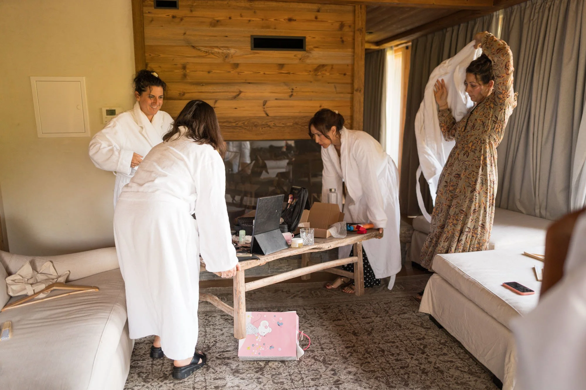 Cinq femmes habillées en peignoirs blancs, organisant des affaires dans une pièce avec un mur en bois, une cheminée et un canapé.
