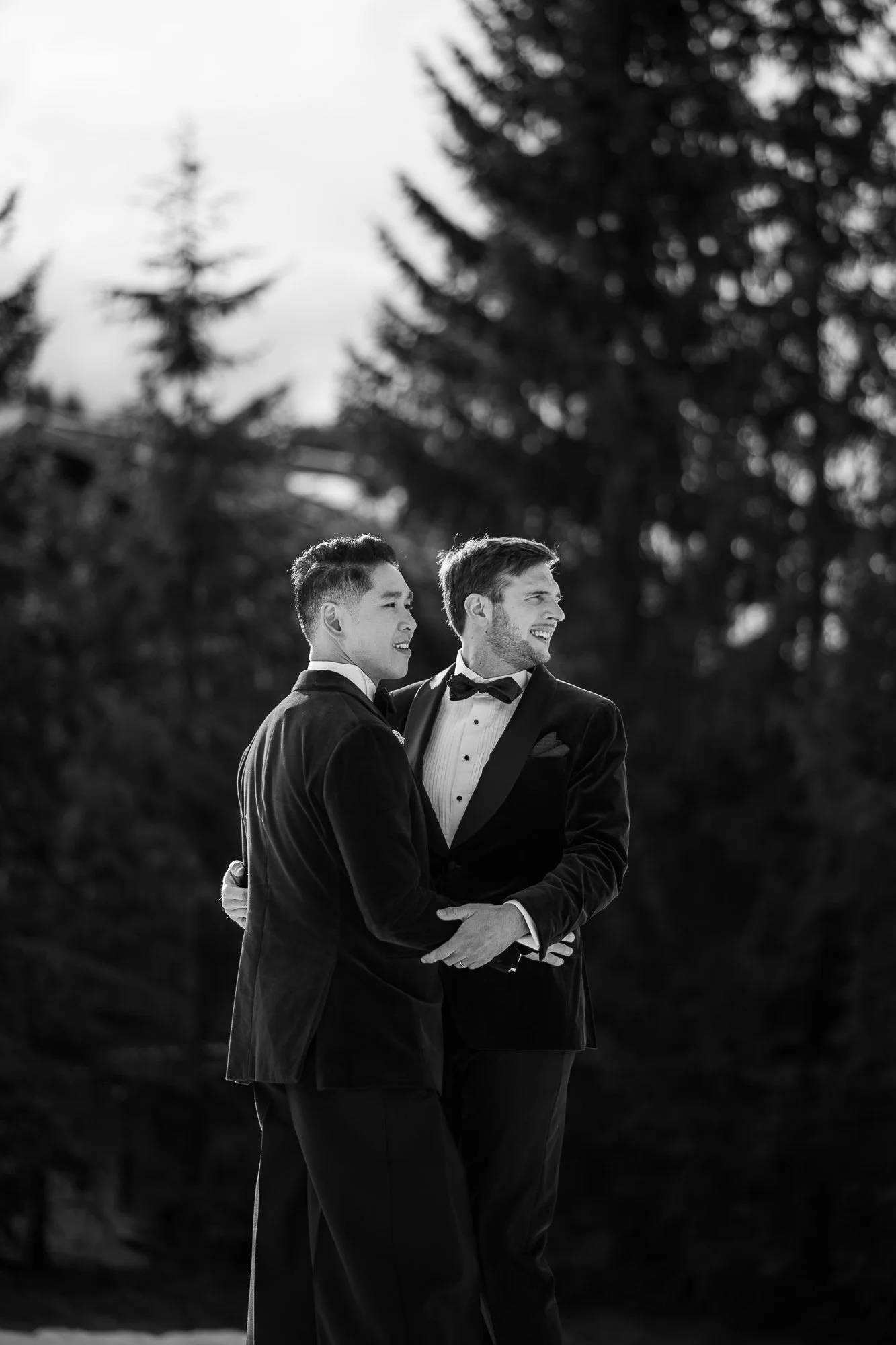 Deux hommes en tuxedo s'embrassent et se regardent avec bonheur dans un environnement naturel, en noir et blanc.