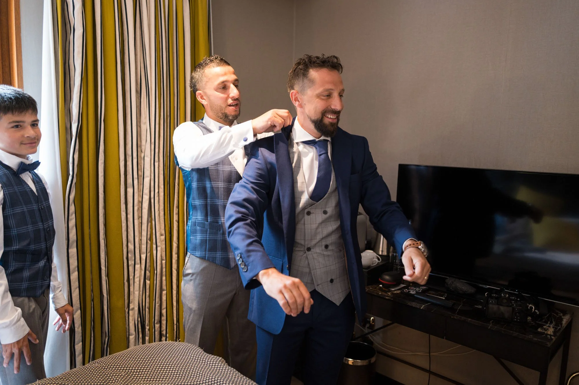Homme en costume bleu en train de se faire aider à enfiler sa veste par un autre homme, deux jeunes garçons habillés en costume sont à côté, dans une pièce avec des rideaux jaunes et vertes.