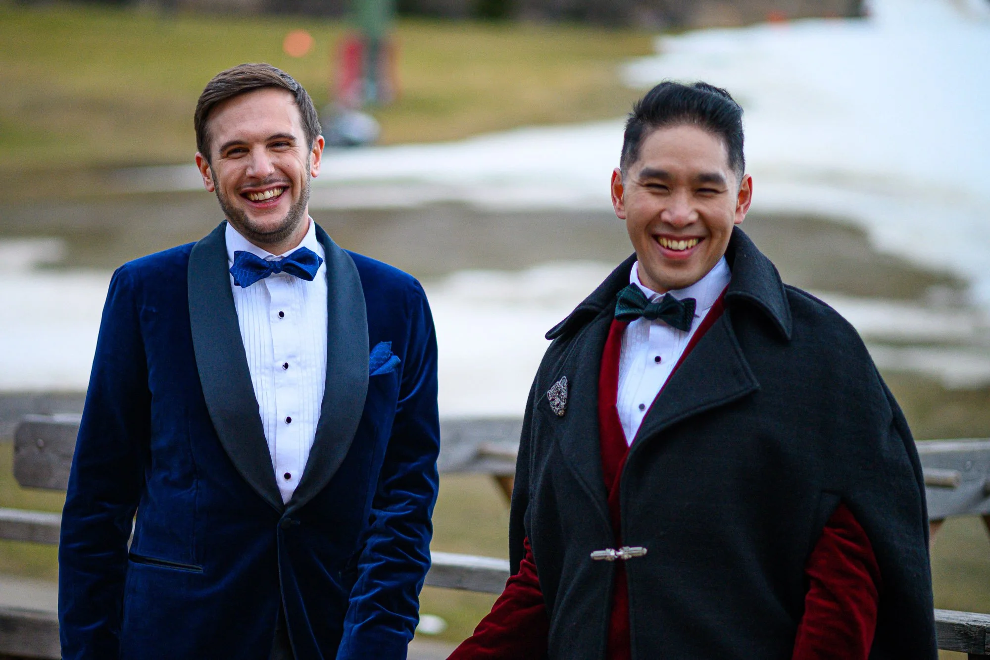 Deux hommes en vêtements formels sourient, l'un en smoking bleu avec nœud papillon et l'autre avec une veste noire et un nœud papillon, dans un parc près de l'eau.