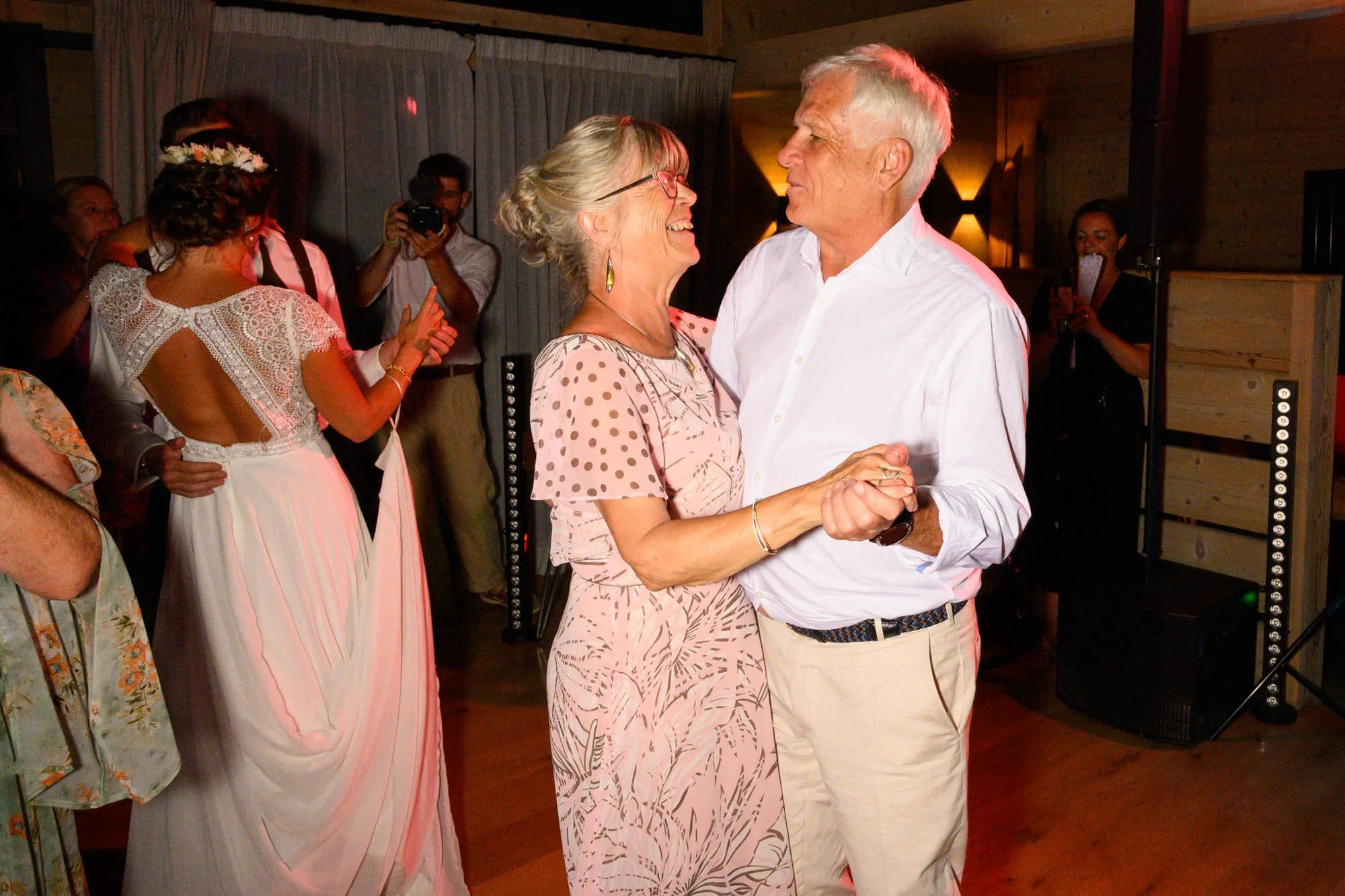 Un couple âgé danse ensemble à une fête, souriant, avec d'autres personnes en arrière-plan, dont une femme en robe blanche avec dos nu, et une personne prenant une photo, dans une ambiance chaleureuse 