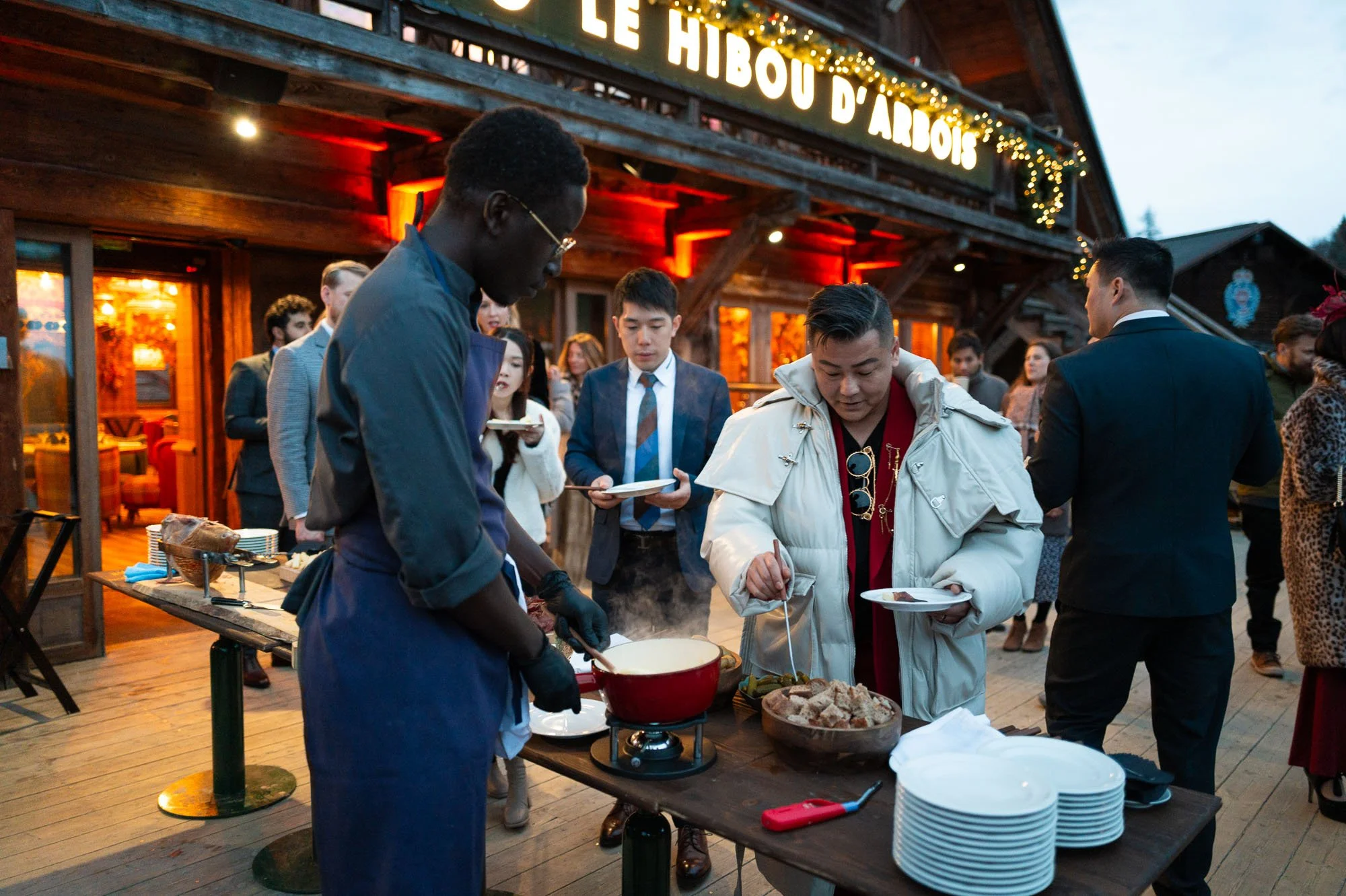 Groupe de personnes à une fête en plein air, servant de la nourriture chaude sur une table en bois avec un fond d'enseigne lumineuse et une cabane en bois décorée de guirlandes lumineuses
