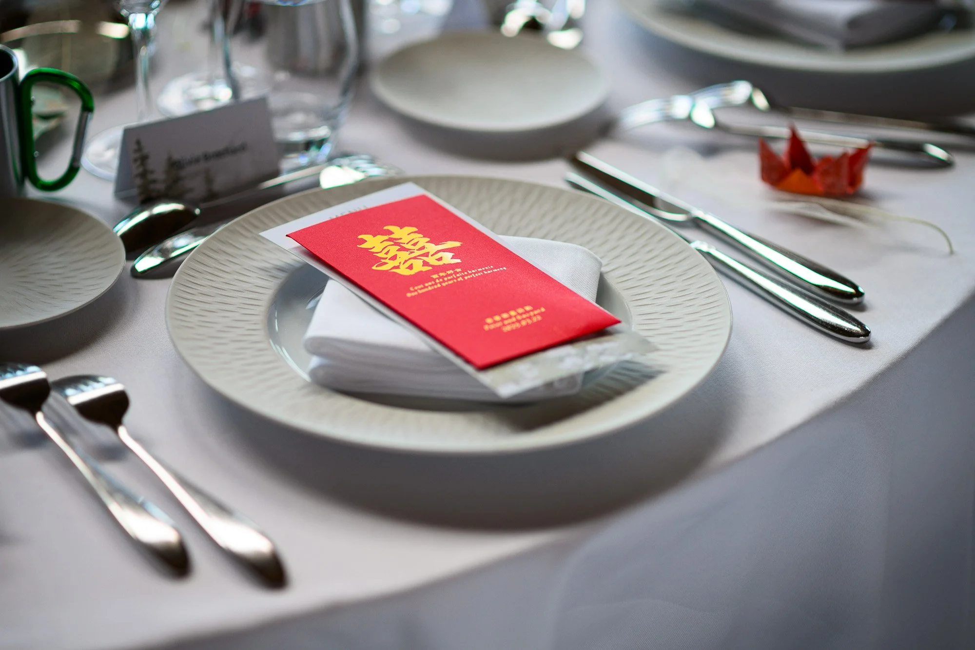 Table dressée pour un banquet avec une assiette, une serviette blanche, une carte rouge et jaune, ciseaux, fourchette, couteau et cuillère en métal brillant, et un décor en forme de navet en papier rouge et orange.