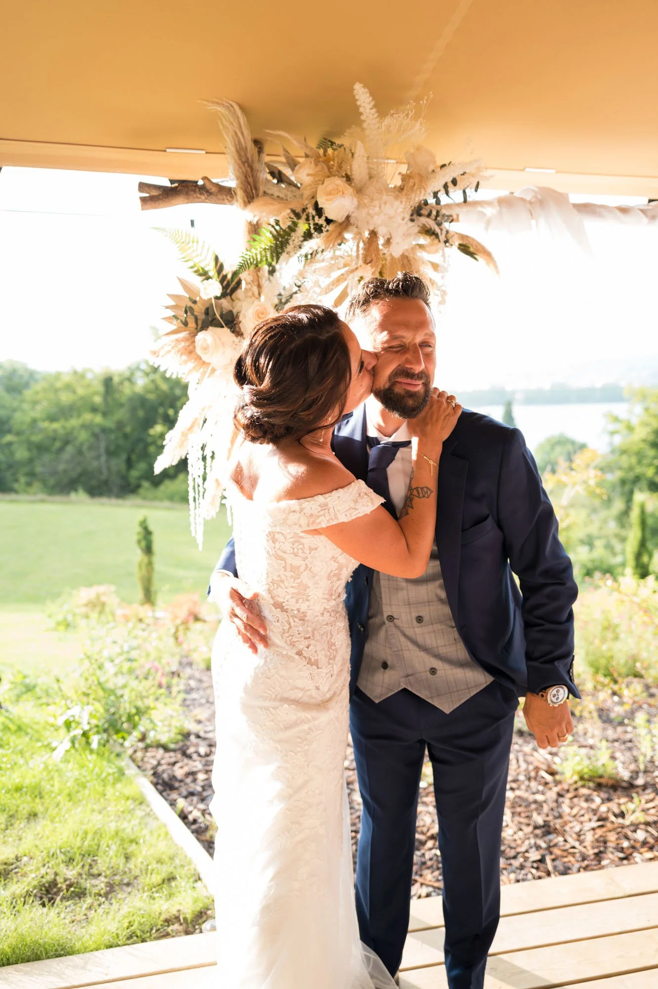 Une femme en robe de mariée embrasse un homme en costume lors d'un mariage en plein air, sous une décoration florale.