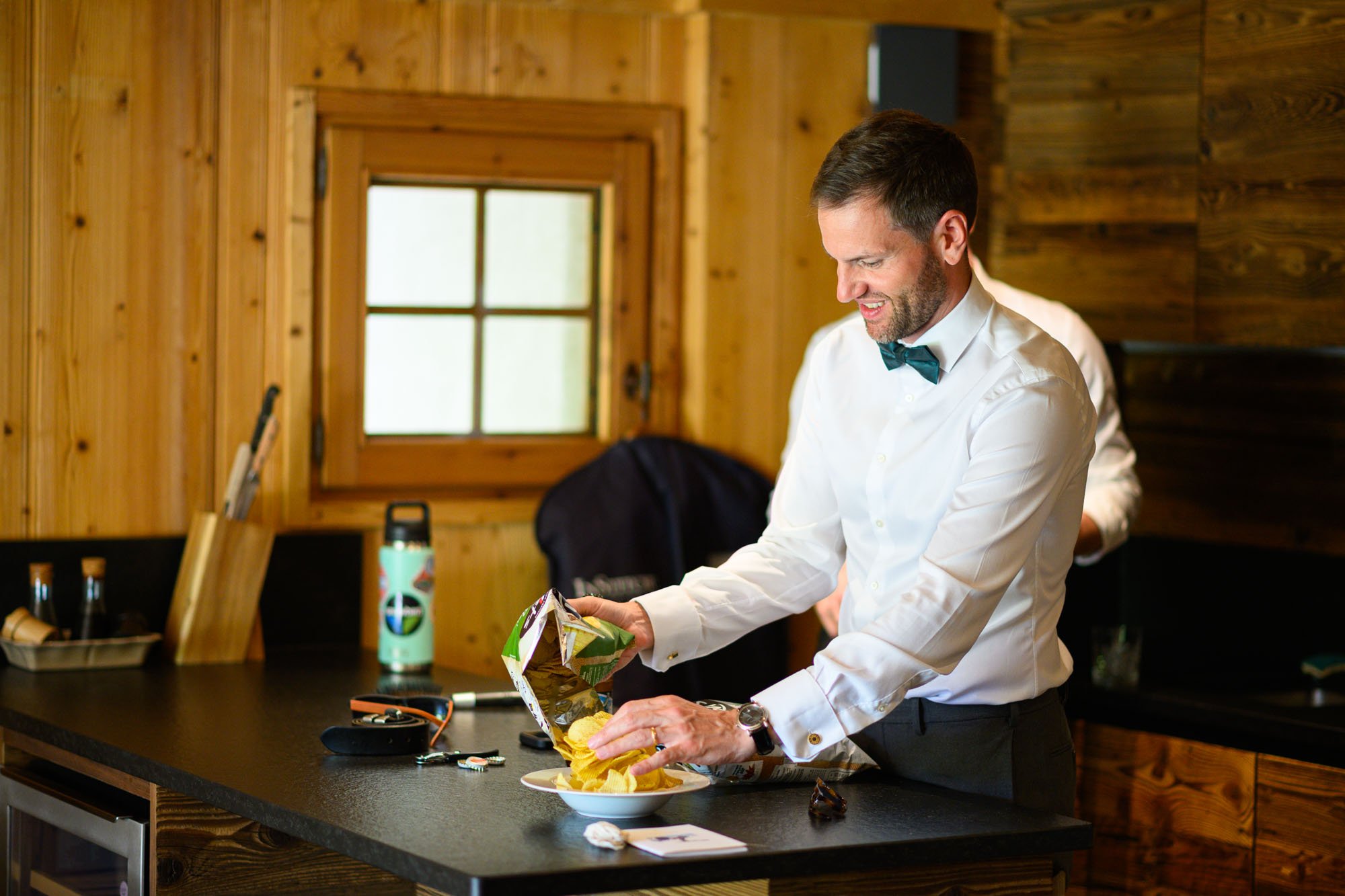 Un homme en chemise blanche et nœud papillon foncé ouvre un paquet de chips dans une cuisine en bois, souriant.