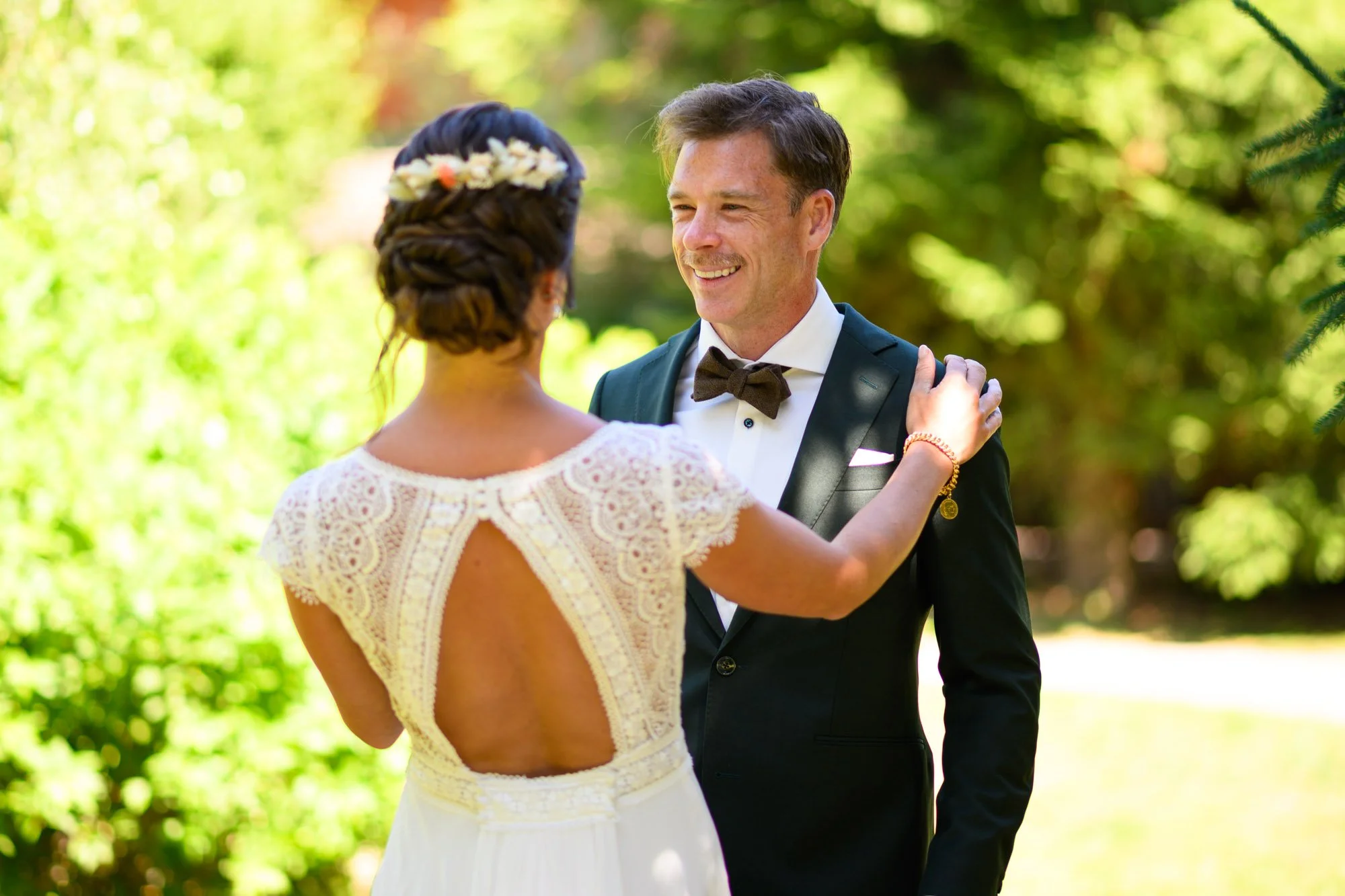 Un couple lors d'un mariage en plein air, la femme en robe blanche avec un dos nu et le homme en smoking avec nœud papillon, échangeant un regard heureux.