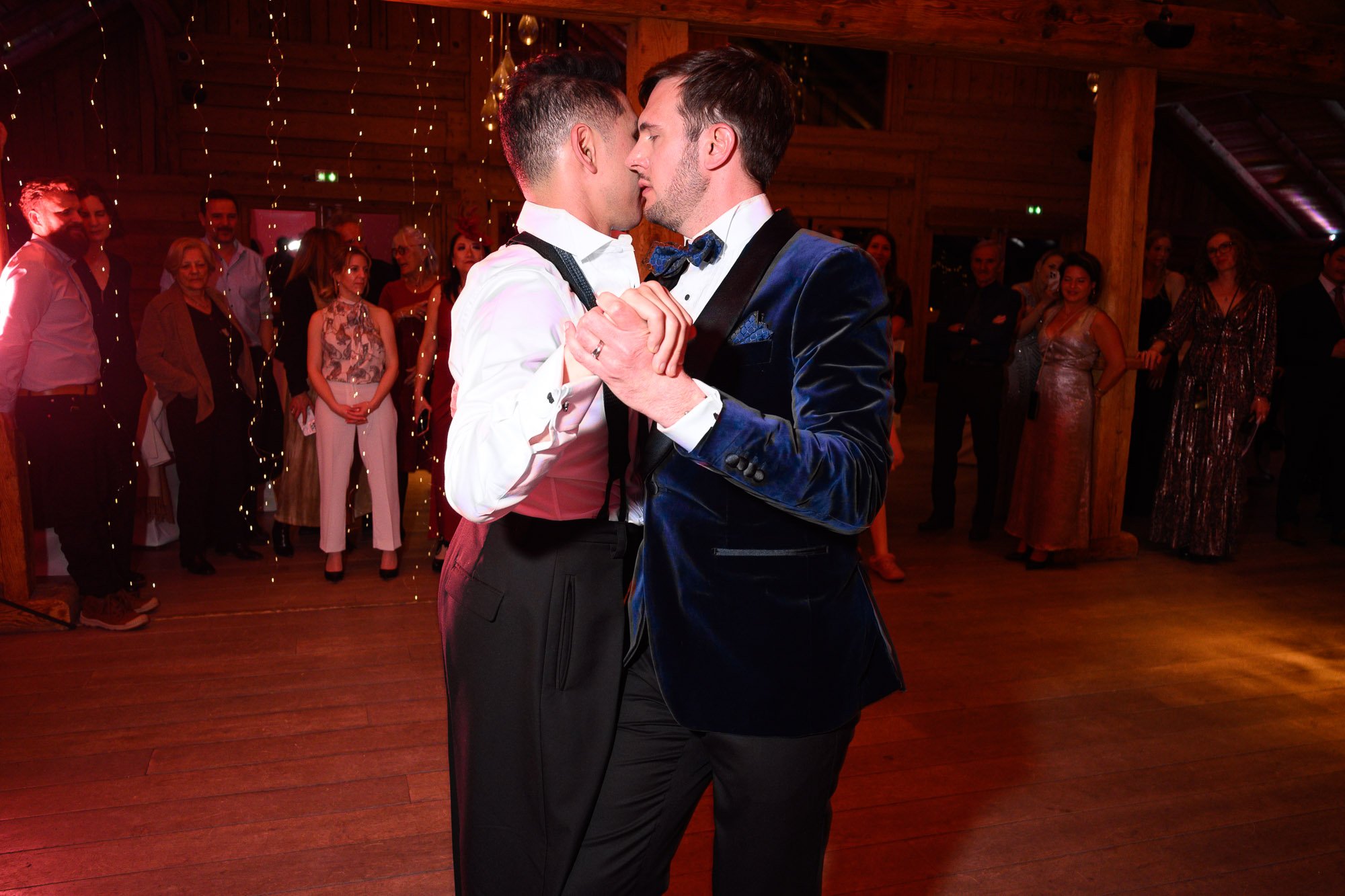 Deux hommes dansent en tête-à-tête lors d'une cérémonie ou d'une fête, entourés de plusieurs personnes qui regardent. La scène se déroule dans un lieu avec un décor en bois chaud et des lumières suspendues.