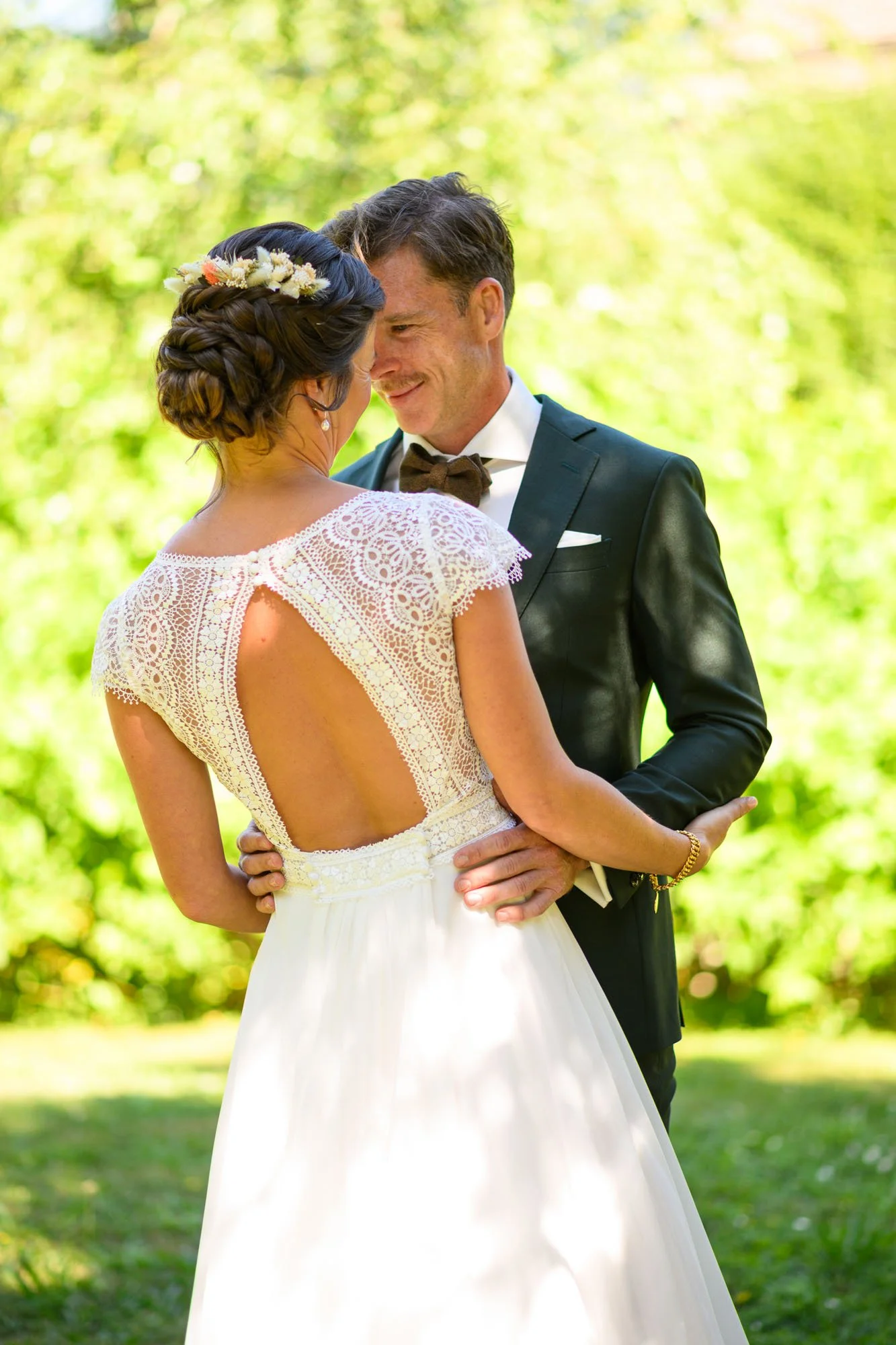 Un couple de mariage danse en plein air, la femme porte une robe blanche avec un dos ouvert en dentelle, et le homme porte un costume noir avec un nœud papillon, dans un décor bucolique avec des arbres verts.