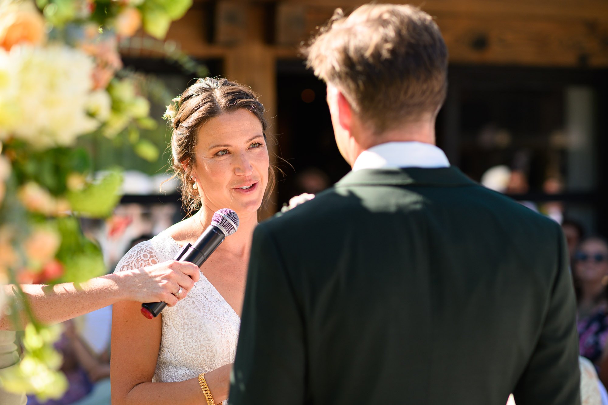 Un mariage en plein air avec une femme tenant un microphone, probablement en train de faire un discours devant un homme en costume, avec des invités en arrière-plan.