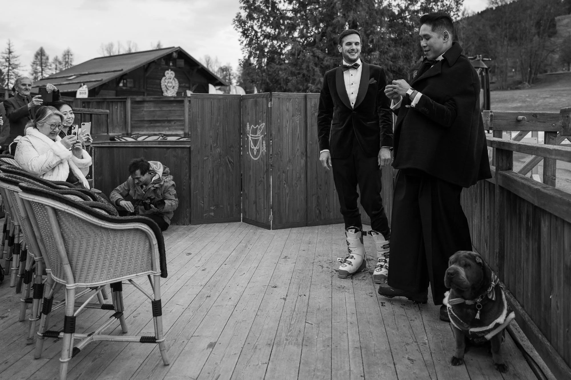 Deux hommes en costumes de mariage, l'un en bas ski, prennent une photo ou une vidéo, entourés par des spectateurs, dont une femme et un homme avec un appareil photo, et un chien avec un vêtement. La scène se déroule sur une terrasse en bois en plein