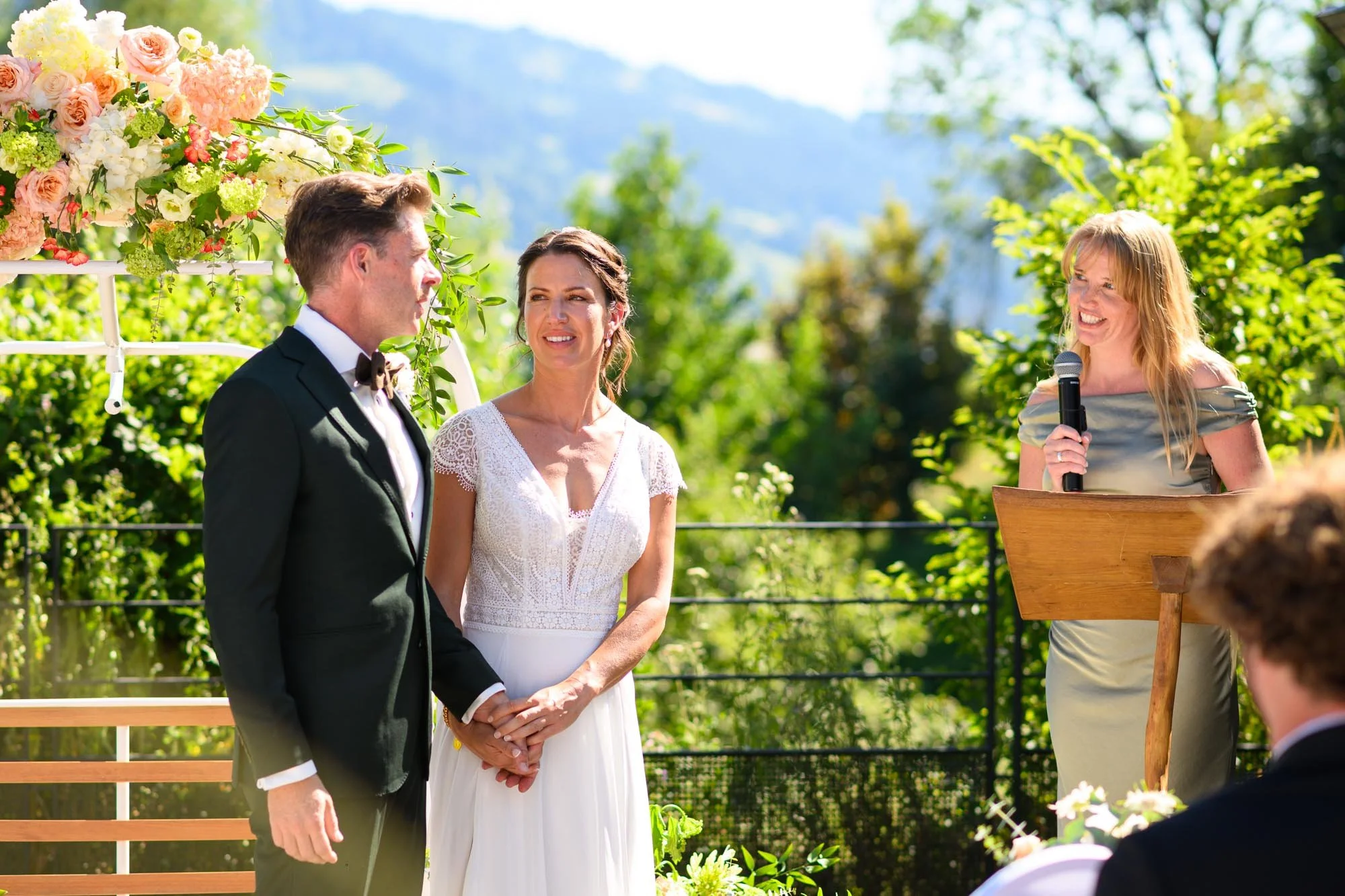 Un couple marié lors d'une cérémonie de mariage dehors, avec une femme qui parle au micro à droite, en arrière-plan et entouré de nature verte et de fleurs.
