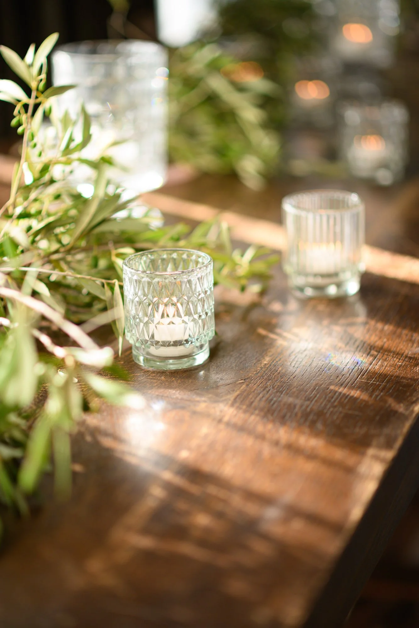 Coupes en verre avec bougies à l'intérieur, décorées avec des branches vertes, disposées sur une table en bois naturel, avec un éclairage doux et naturel.