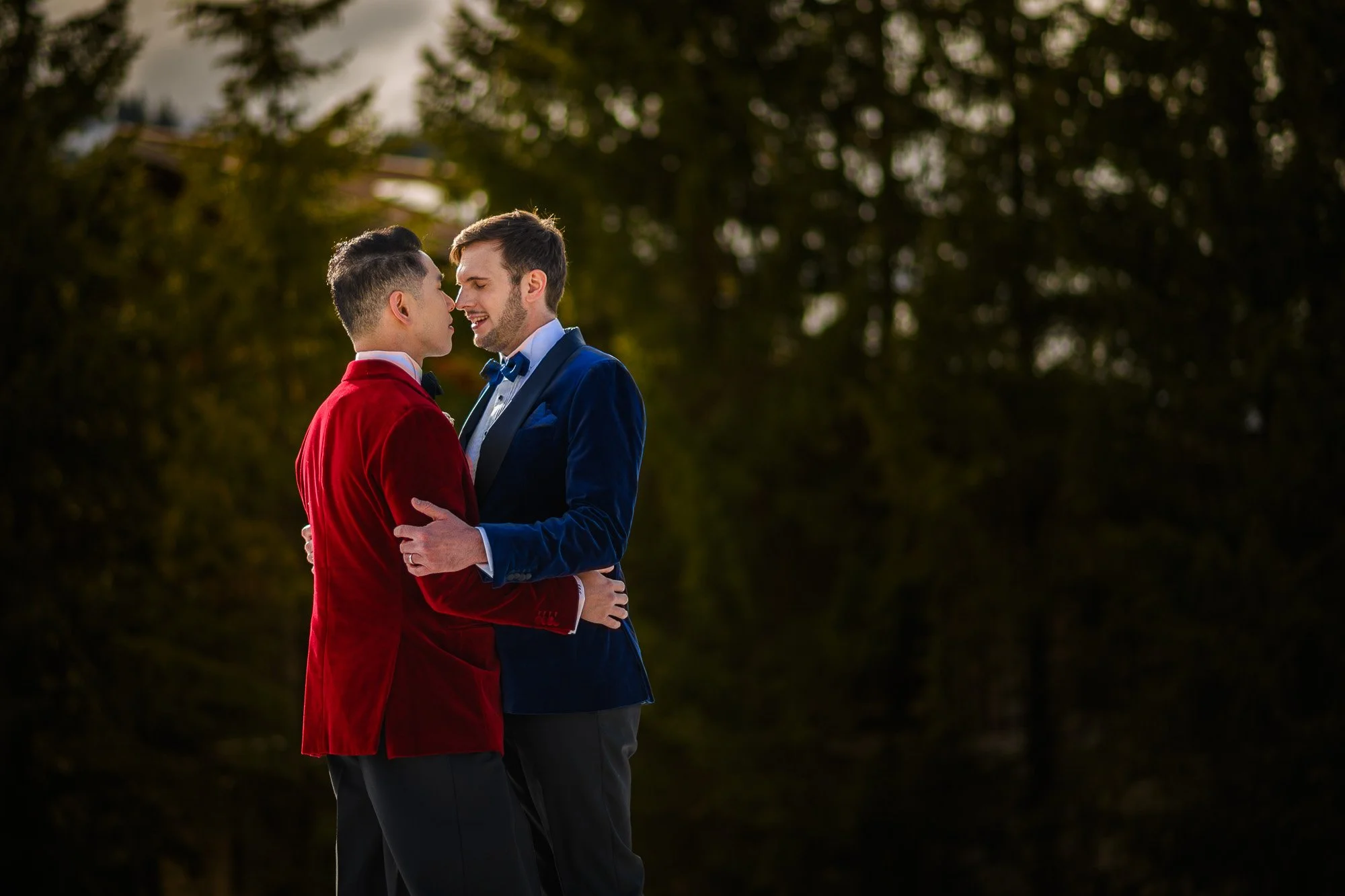 Deux hommes habillés en smokings colorés, un en rouge et l'autre en bleu, se tenant proches face à face dans un parc avec des arbres en arrière-plan.