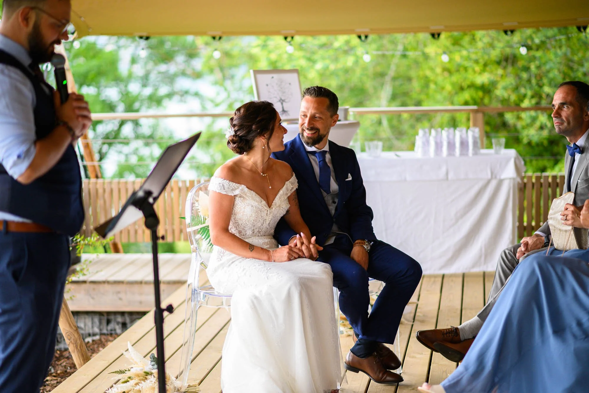 Un mariage en plein air avec un couple assis lors de la cérémonie, la femme en robe blanche et l'homme en costume bleu, qui se tiennent la main et se regardent avec bonheur. Un officiant se tient à gauche, et des invités sont assis, assistant à la cé
