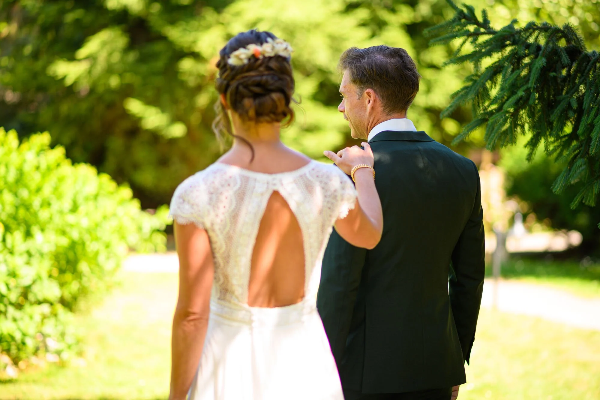 Un couple en mariage, une femme en robe de mariée blanche avec un dos ouvert et un homme en costume noir, se tenant dans un cadre verdoyant lors d'une journée ensoleillée.