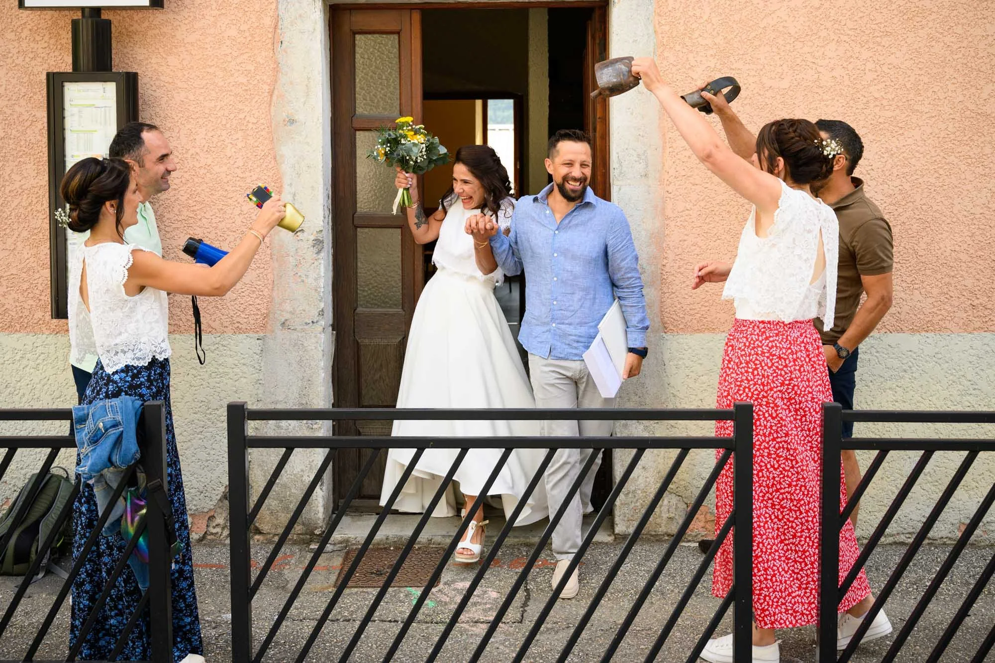 Un groupe de personnes célébrant devant une porte, avec une femme en robe blanche tenant un bouquet, entourée de personnes qui semblent festives et joyeuses.
