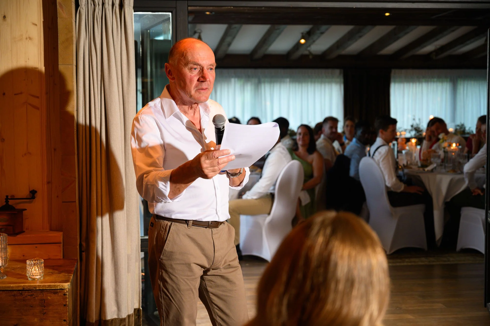 Un homme âgé avec une chemise blanche tient un micro et un papier, parlant devant un groupe assis lors d'un événement ou d'une réception dans une salle décorée.
