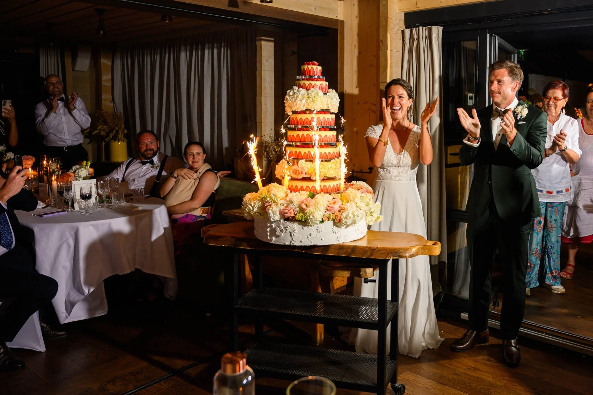 Un couple de mariés en train de couper un grand gâteau nuptial décoré de fleurs et de sparklers, entouré d'invités qui applaudissent et prennent des photos dans une réception de mariage chaleureuse et festive.