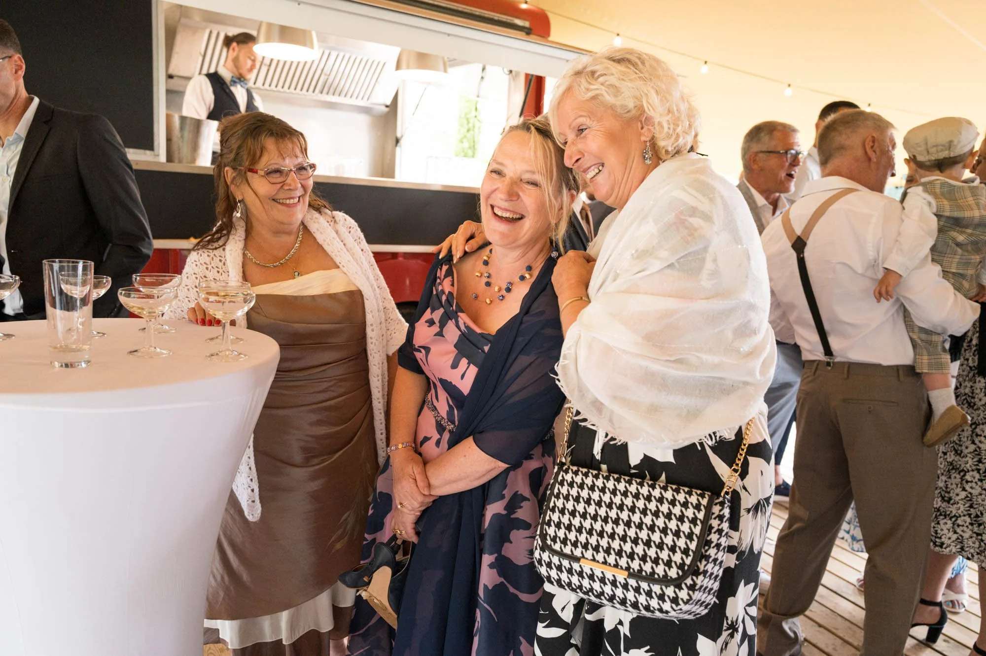 Des femmes souriantes en tenues élégantes, discutant lors d'une fête ou un mariage, avec un groupe de personnes en arrière-plan.