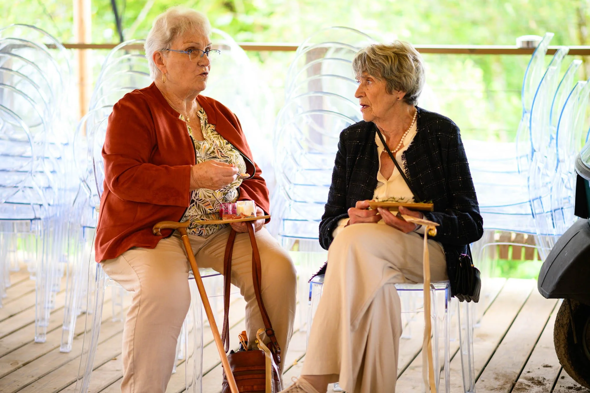 Deux femmes âgées discutent assises sur des chaises en plastique transparent, en extérieur, avec un fond de verdure.