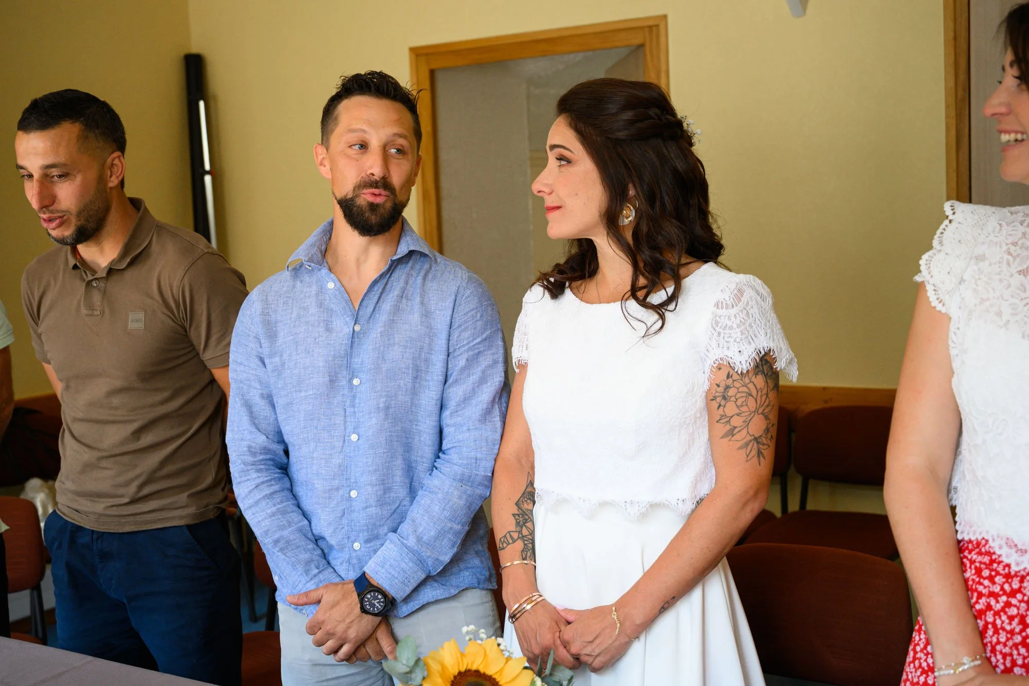 Un groupe de personnes lors d'une cérémonie, avec une femme portant une robe blanche avec dentelle, discutant avec un homme en chemise bleue, autres personnes visibles en arrière-plan.