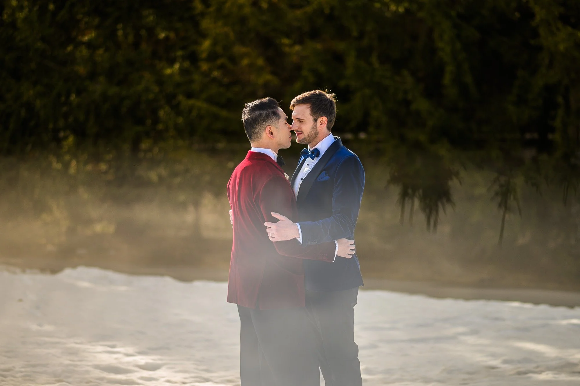 Deux hommes en costumes élégants s'embrassent tendrement sur une plage, avec une forêt en arrière-plan et une brume légère au sol.
