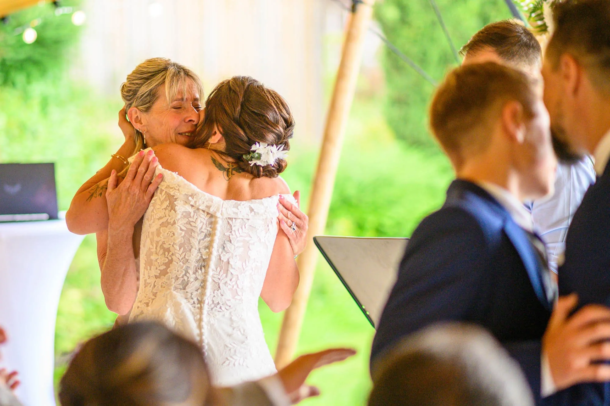 Deux femmes âgées s'embrassent lors d'un mariage, avec des invités autour d'elles, en intérieur avec un décor naturel en arrière-plan.