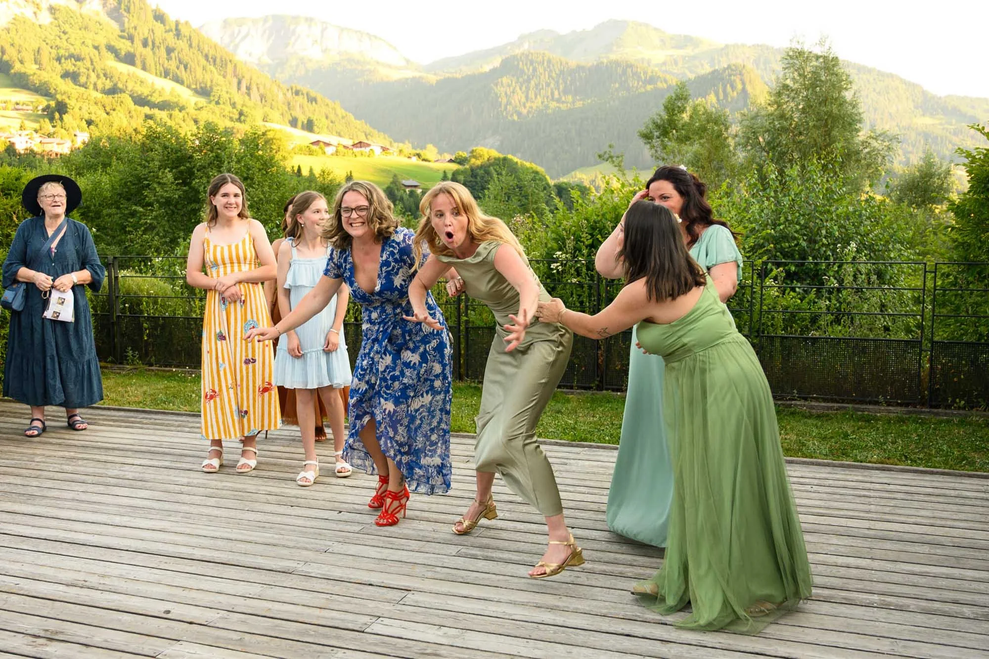 Groupe de femmes en robes colorées, certaines riant et d'autres regardant, sur une terrasse en bois avec un paysage de montagnes et de verdure en arrière-plan.