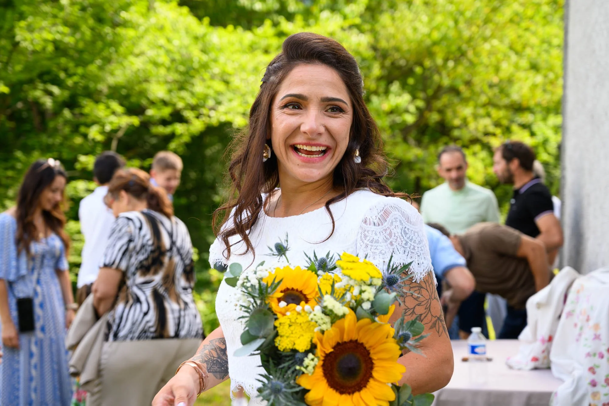 Une femme souriante dans un jardin lors d'une fête ou d'un mariage, tenant un bouquet de tournesols et autres fleurs, avec d'autres personnes en arrière-plan.