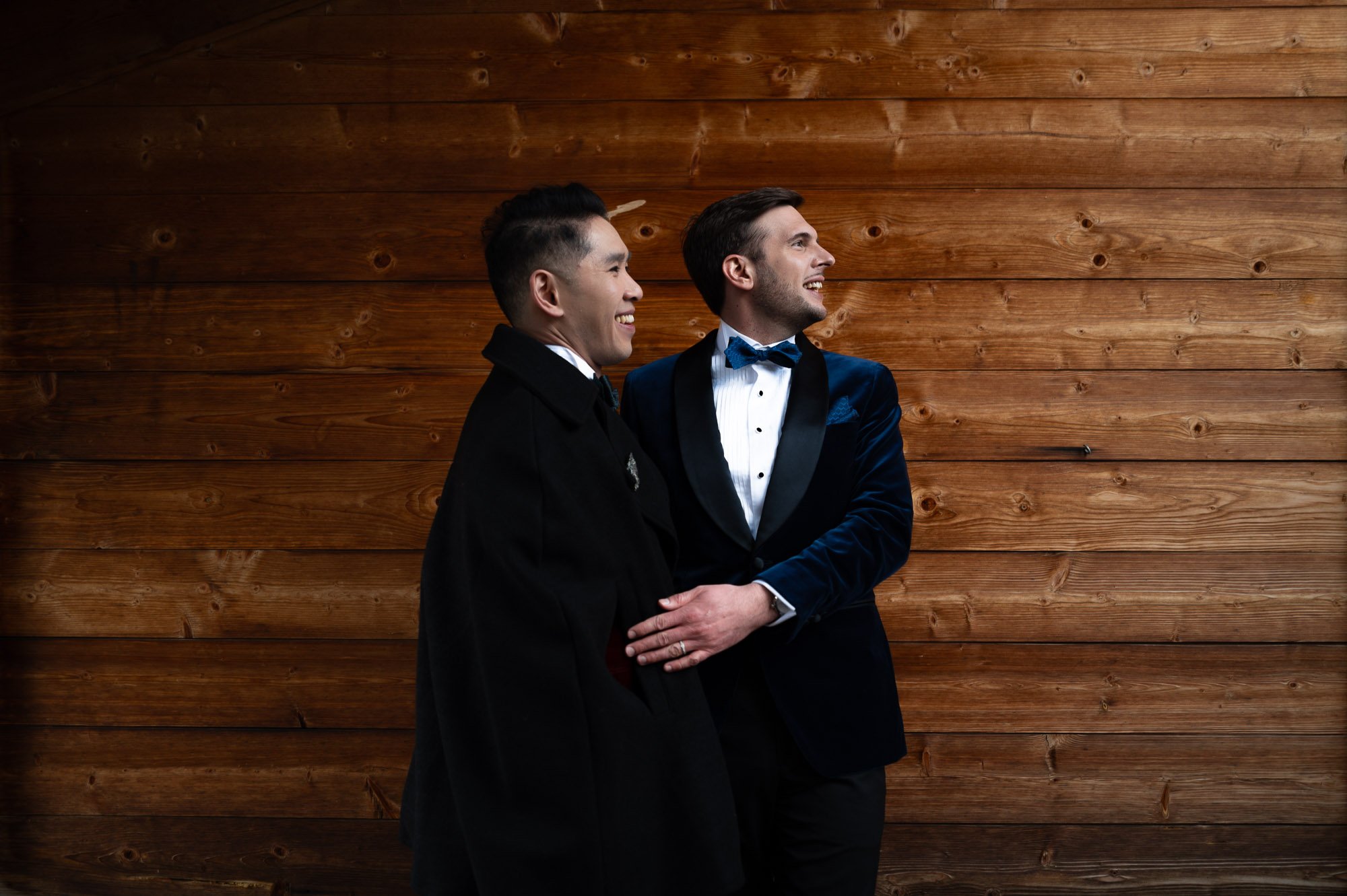 Deux hommes habillés élégamment se tenant contre un mur en bois, souriant et discutant ensemble.