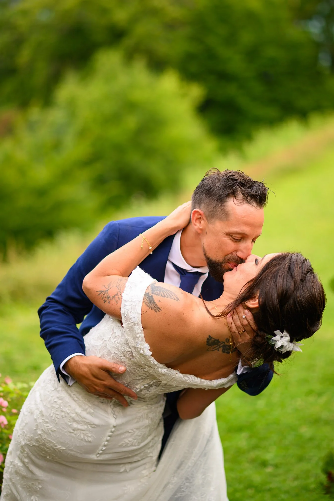 Un couple marié s'embrassant dans un jardin en plein air, la femme porte une robe de mariage blanche avec des tatouages visibles, et l'homme porte un costume bleu marin.