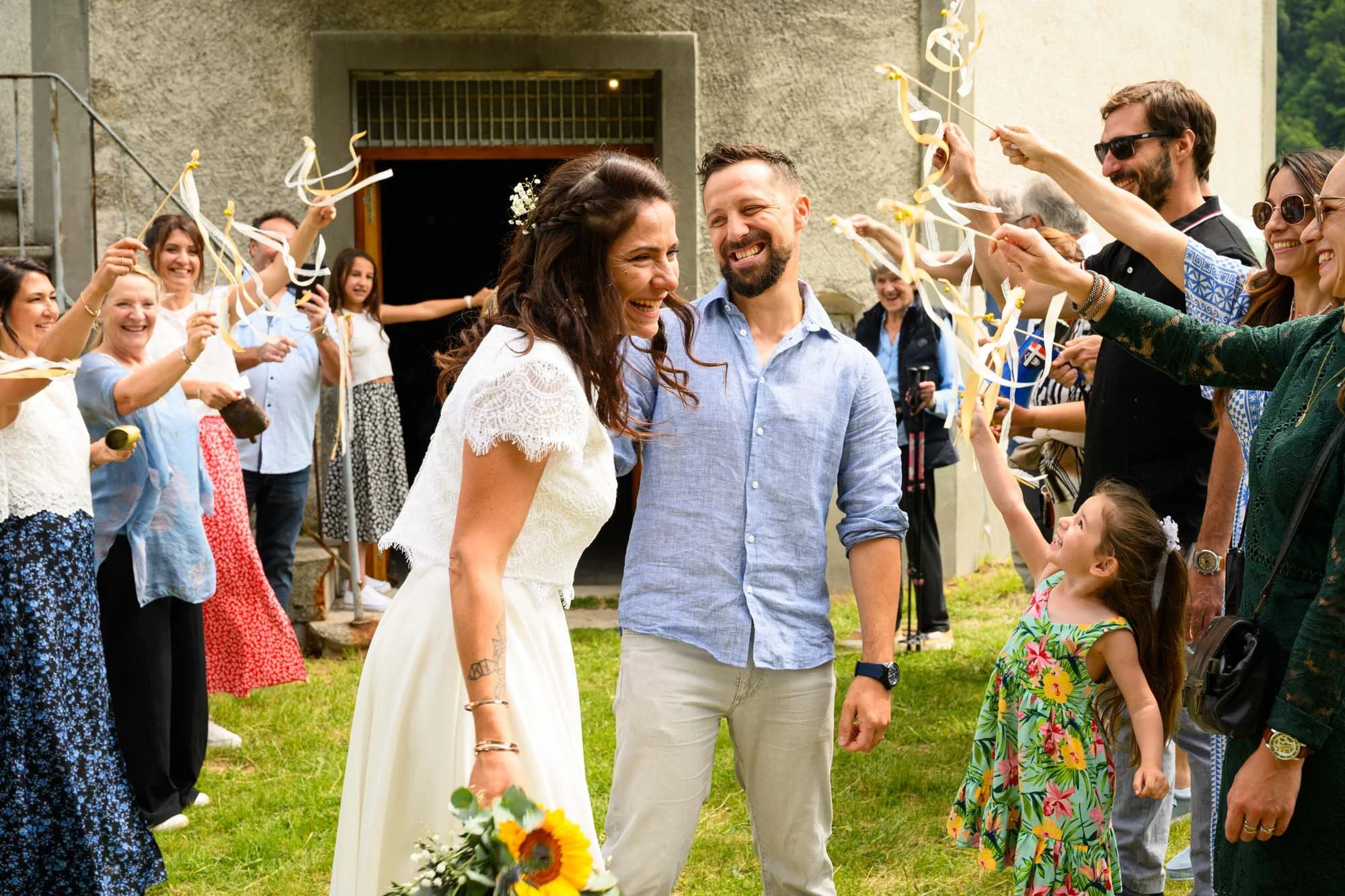 Un couple de mariés souriants entouré de membres de la famille et amis lors d'une célébration de mariage en plein air, avec des invités lançant des rubans et une jeune fille avec un bouquet de fleurs.
