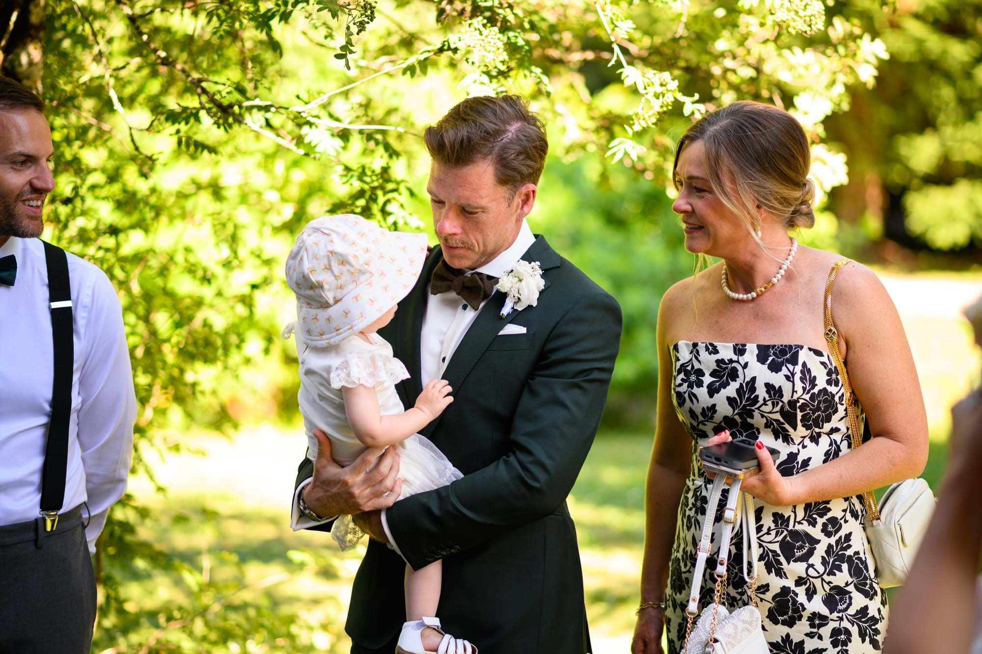 Un homme en costume de mariage tient une petite fille en robe blanche et chapeau à fleurs, lors d'un événement en plein air, avec deux autres adultes autour, sous un arbre vert.