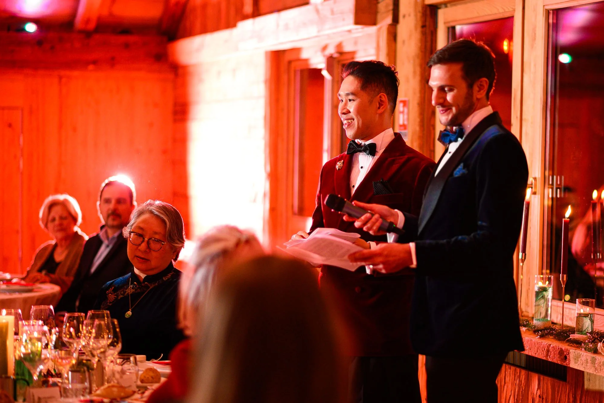 Deux hommes habillés en smoking présentent un discours lors d'un dîner dans une salle avec intérieur en bois, avec des invités à la table et des bougies pour décoration.