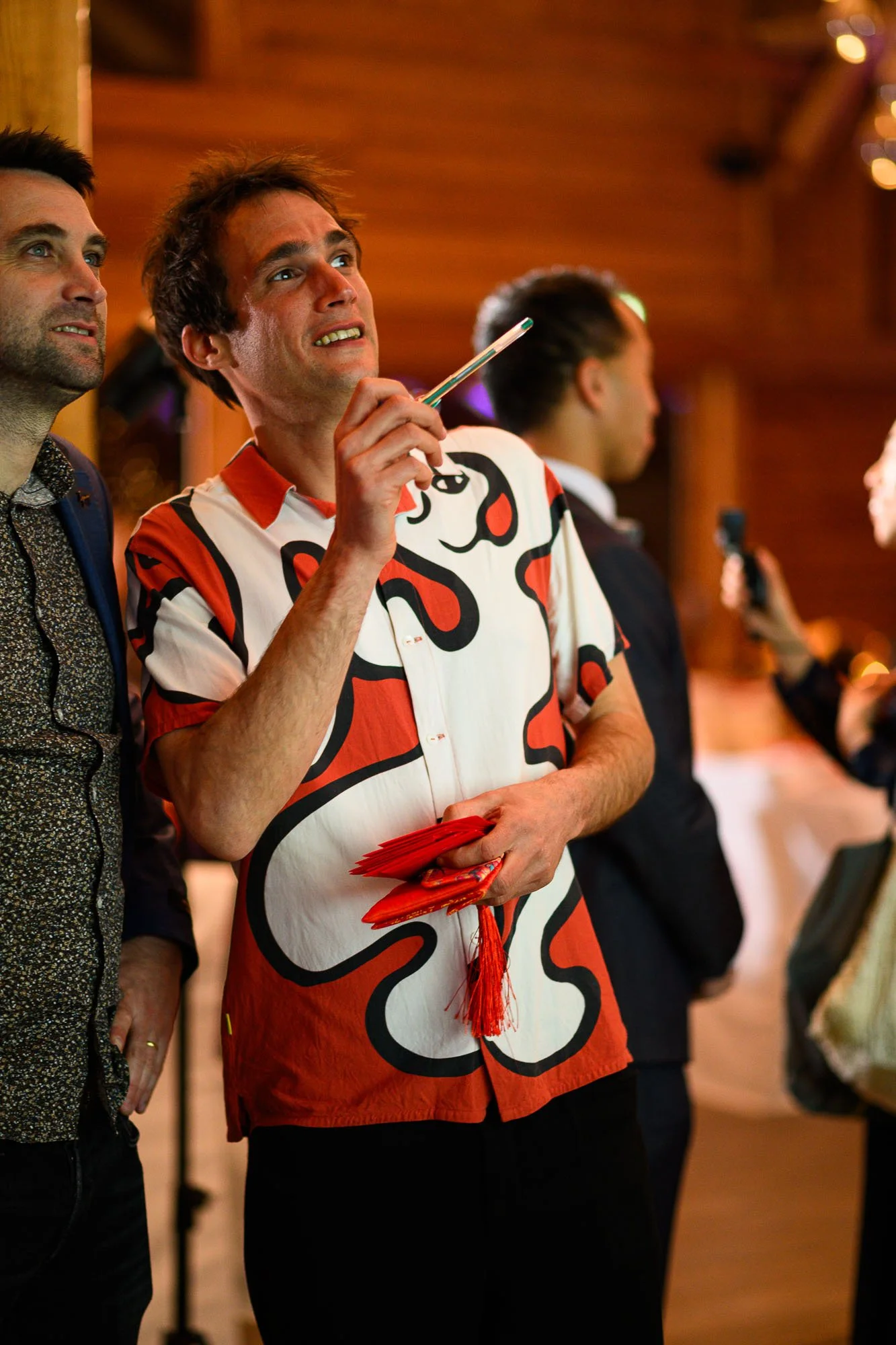 Un homme en chemise colorée avec un motif noir, rouge, blanc tient un bâtonnet ou un pointeur, et semble réfléchir ou s'interroger, entouré d'autres personnes dans un environnement intérieur lumineux.