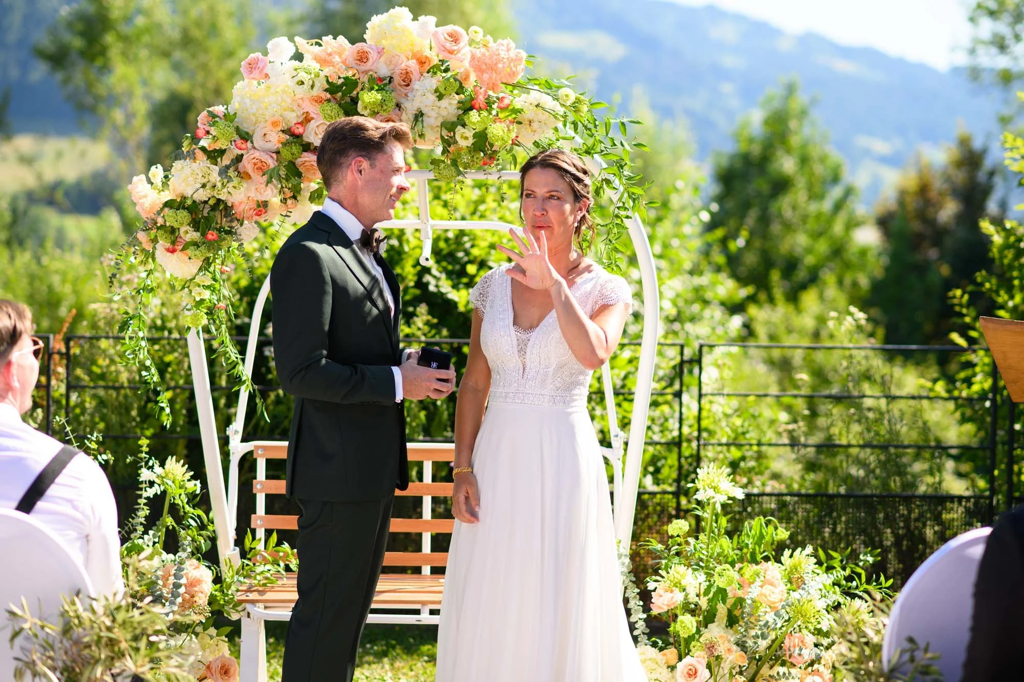 Un couple marié échange des vœux lors d'une cérémonie en plein air avec un décor floral, une arche et un fond verdoyant.