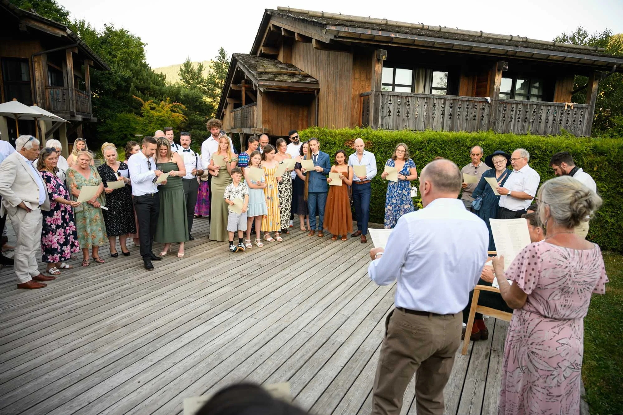 Un groupe de personnes en extérieur, probablement lors d'une cérémonie, chantant en regardant des feuilles, avec des maisons en bois en arrière-plan.