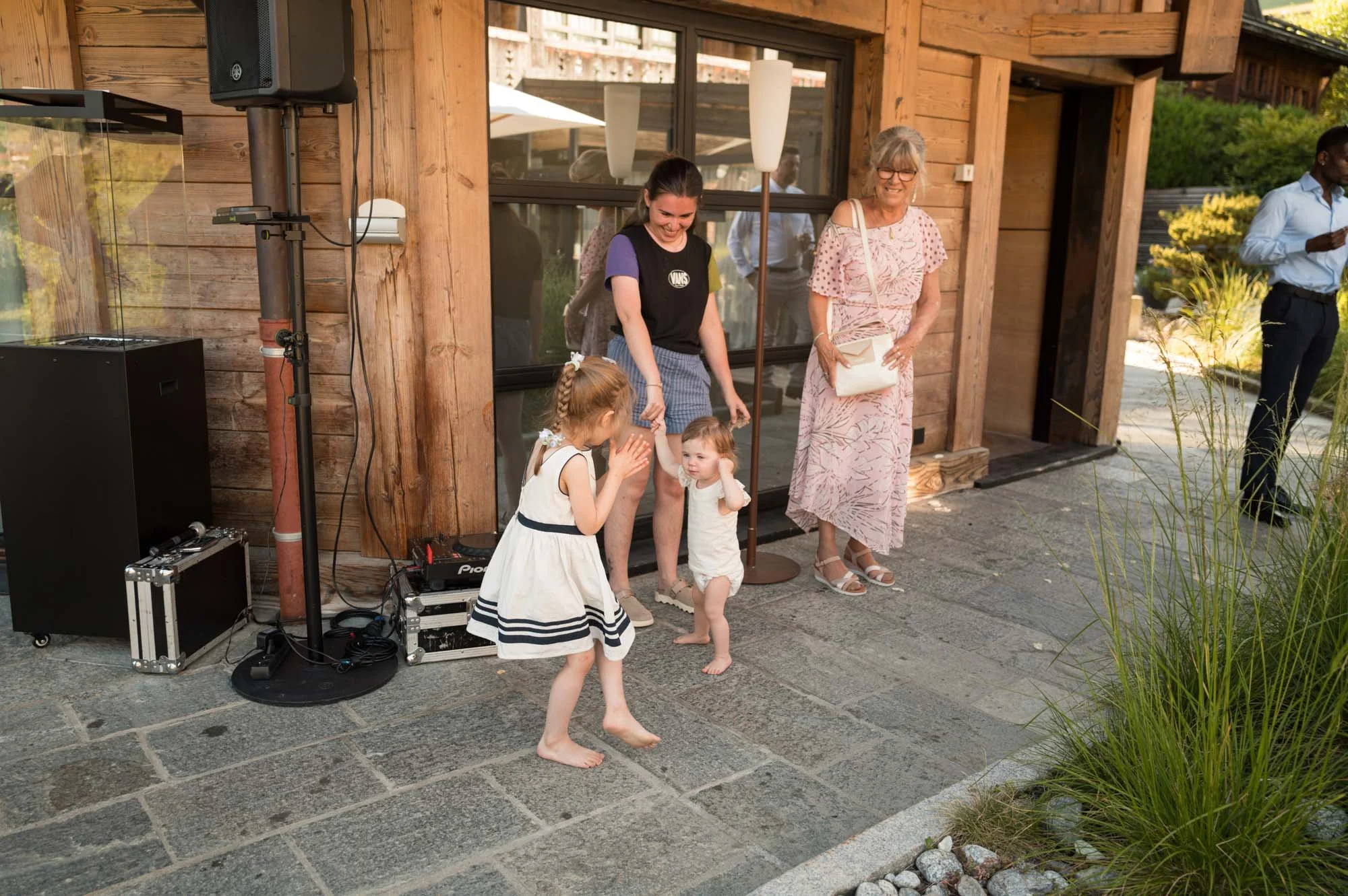 Une femme et une fille dansent, deux autres filles la regardent, devant une maison en bois, avec d'autres personnes en arrière-plan, lors d'une fête extérieure.