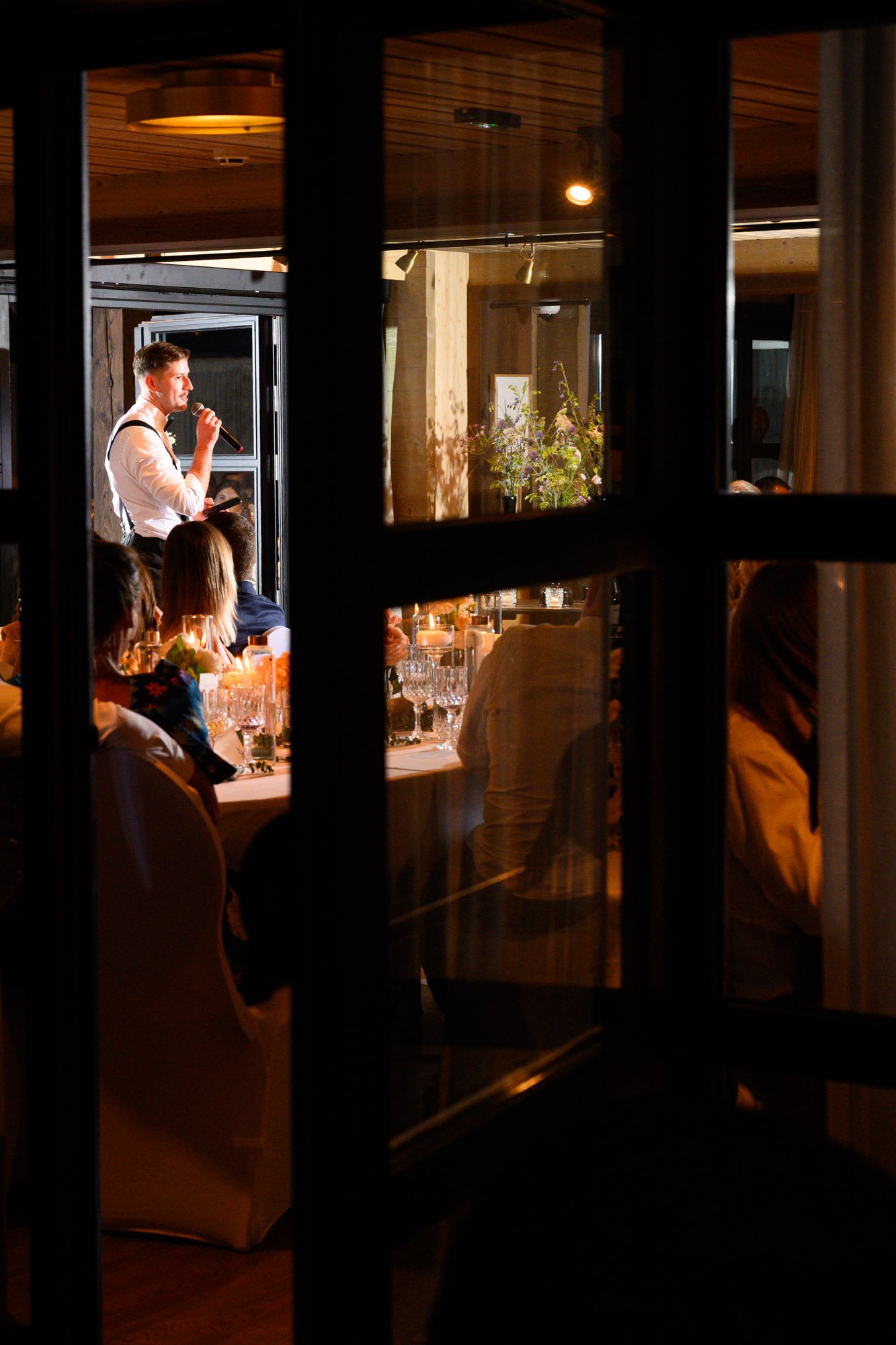 Personne en costume parlant dans un microphone lors d'un dîner, vu à travers une fenêtre par une autre personne.