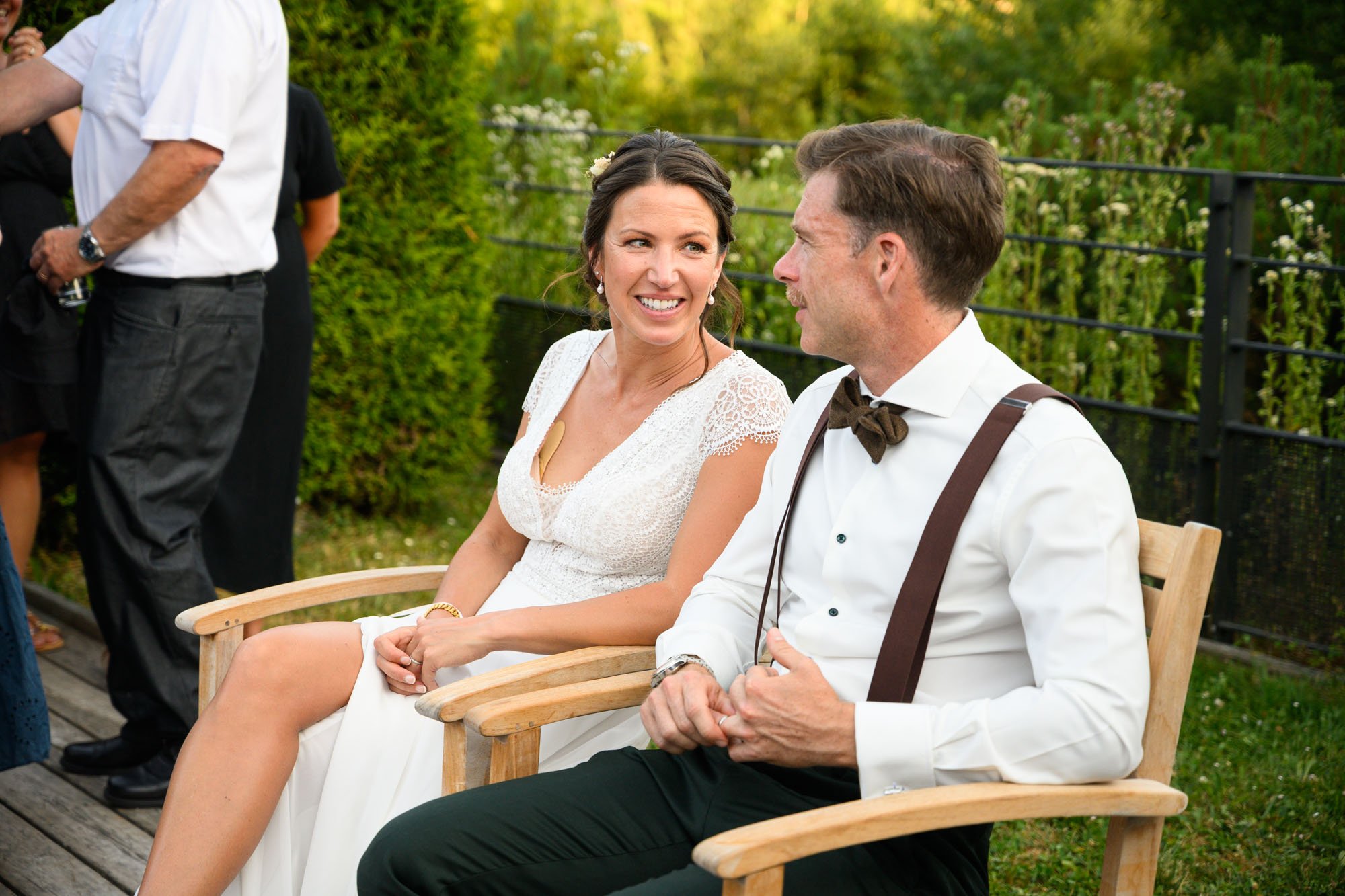 Un couple assis sur des bancs en bois lors d'un mariage, la femme porte une robe blanche et le homme une chemise blanche avec un nœud papillon. Ils semblent discuter et partager un moment heureux dans un jardin.